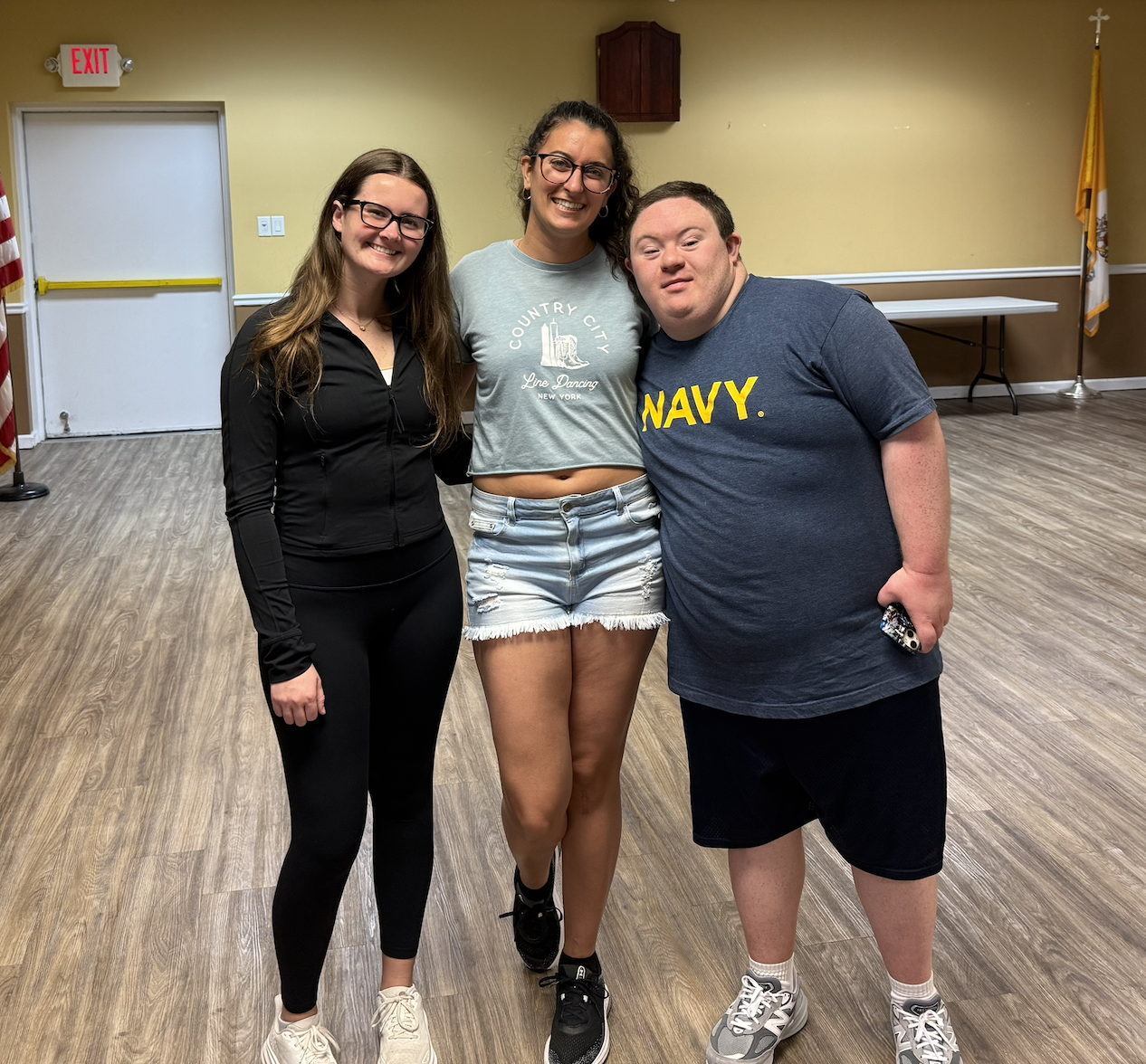 Three people standing together smiling in a room with wood flooring, yellow walls, and American flags in the background.