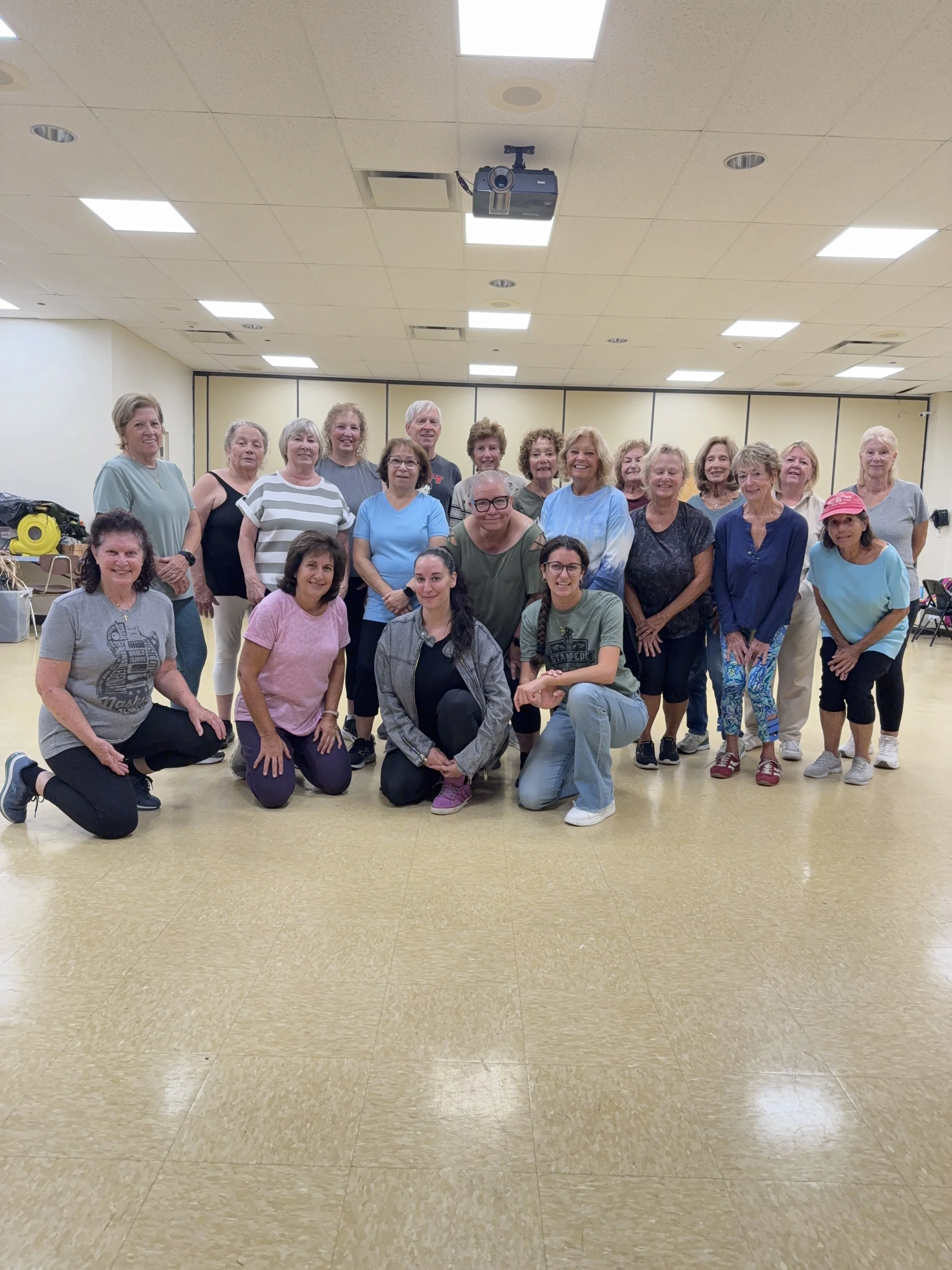 A large group of elderly women and a few men posing for a photo in a community center or dance studio with beige walls and ceiling tiles, and a beige tiled floor.