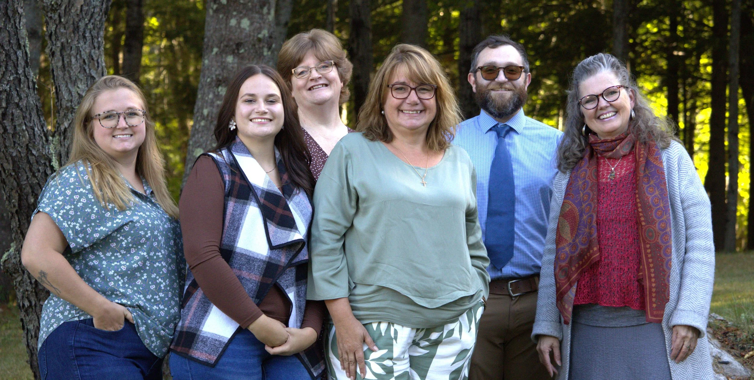 Group of six smiling women and a man standing outdoors in a forest, posing for a photo.