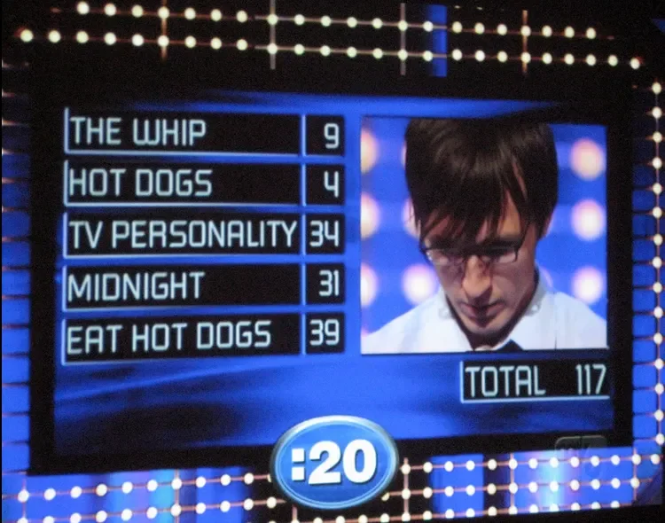 Game show scoreboard displaying category scores: The Whip 9, Hot Dogs 4, TV Personality 34, Midnight 31, Eat Hot Dogs 39, total 117. A timer at 20 seconds and a contestant's face on the right side.
