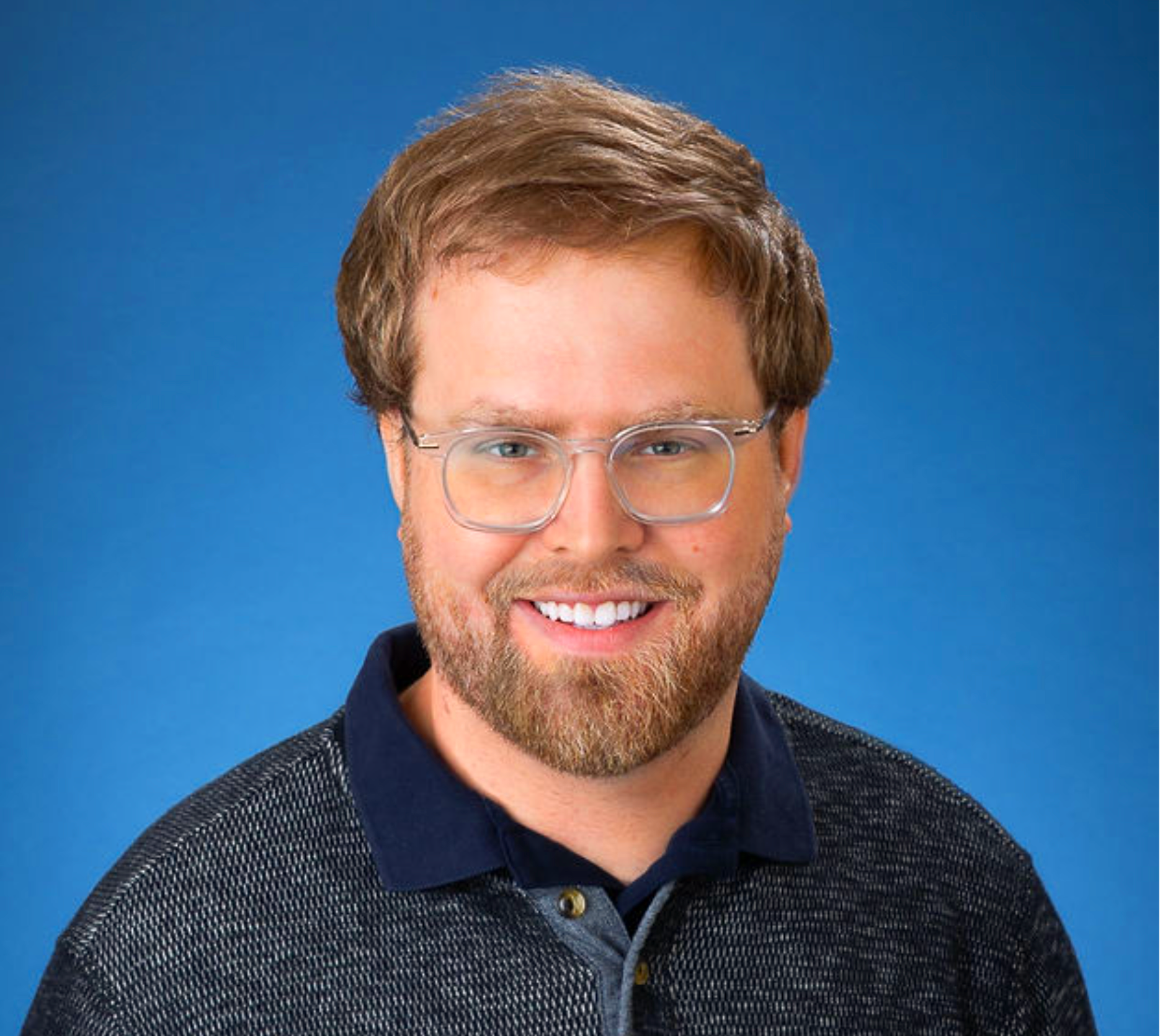 Close-up of a smiling young man with light brown hair, beard, and glasses, wearing a dark collared shirt and jacket, against a blue background.