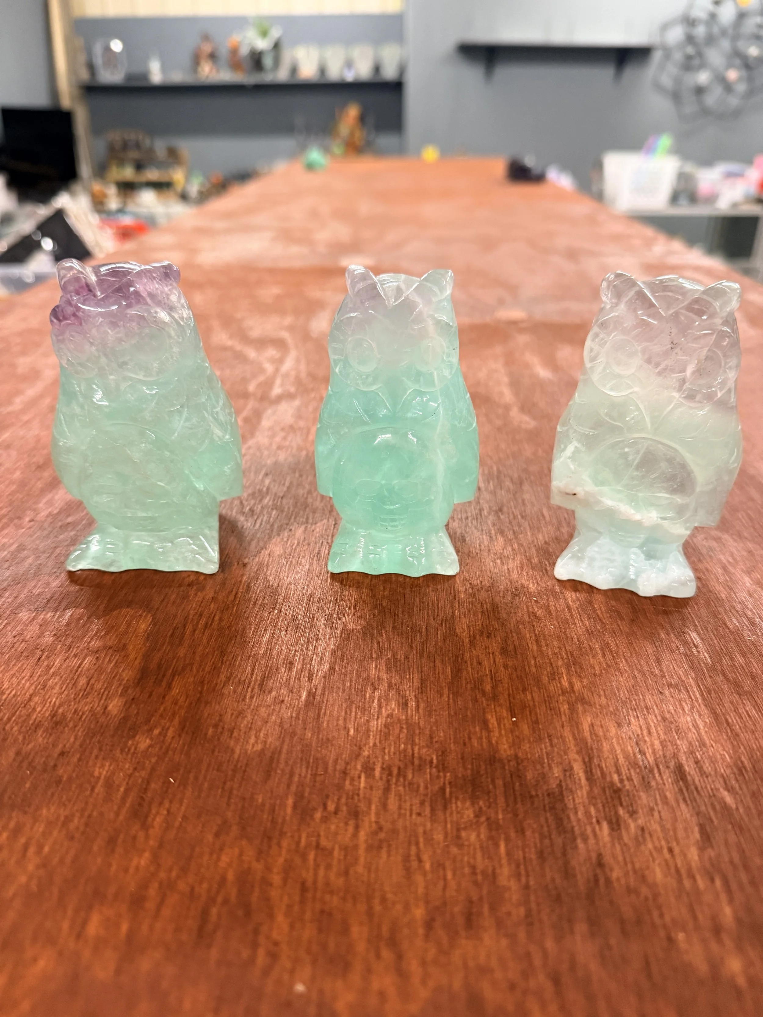 Three carved jade animal figurines, possibly bears, lined up on a wooden table with a blurred background of shelves and decorations.