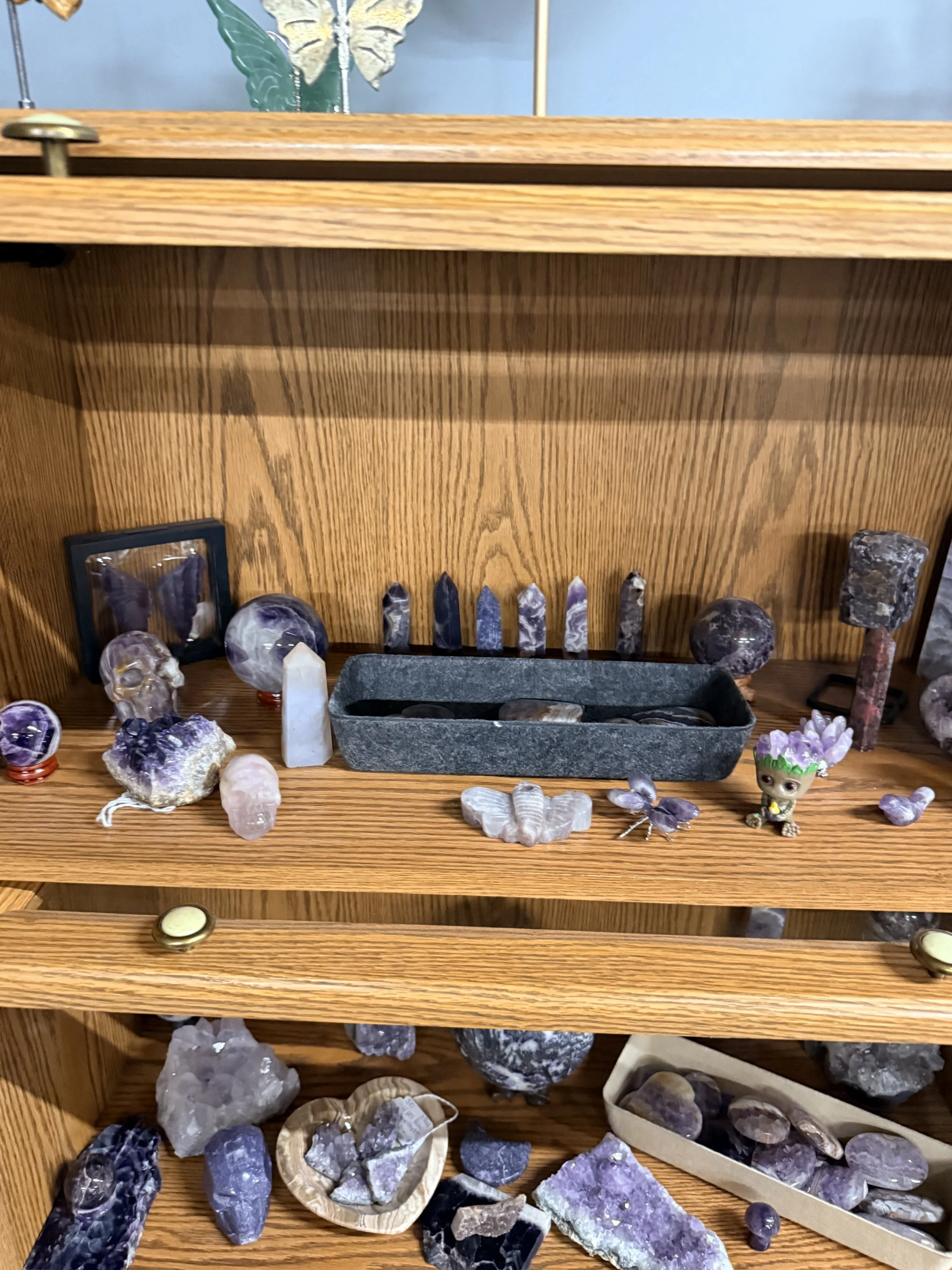 A wooden display shelf with various polished and rough amethyst crystals, some carved into skull shapes, a small figurine with a floral crown, and a black rectangular container holding small amethyst pieces.