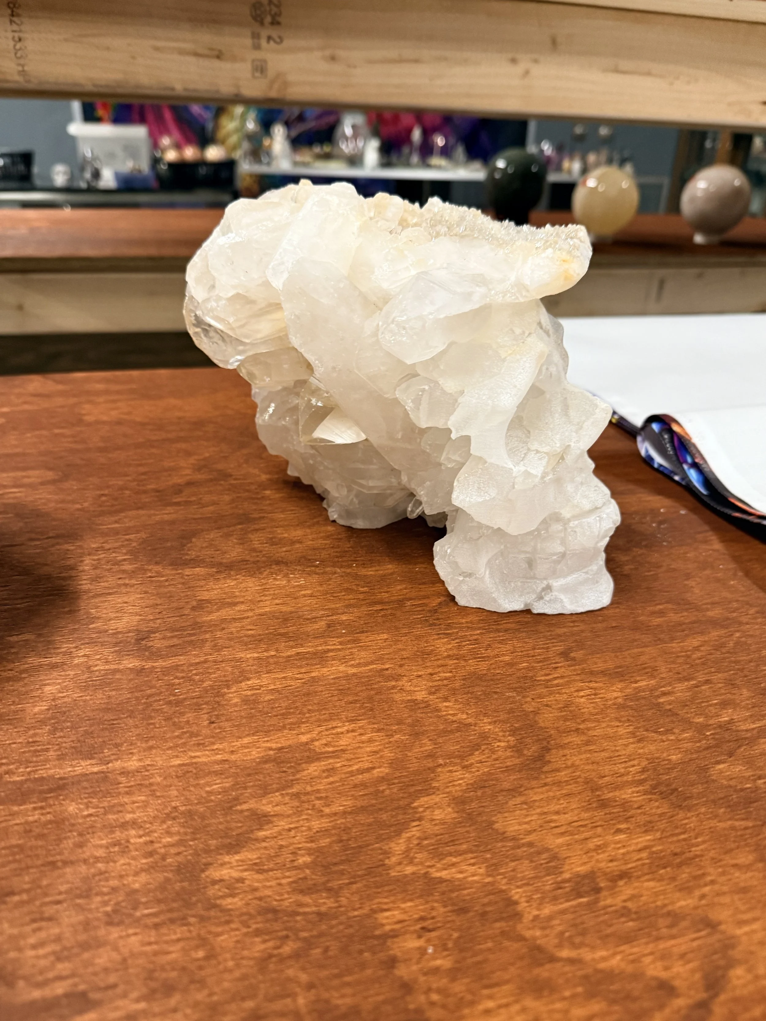 A large white crystal or mineral specimen on a wooden surface with a blurred background showing various objects and colorful items.