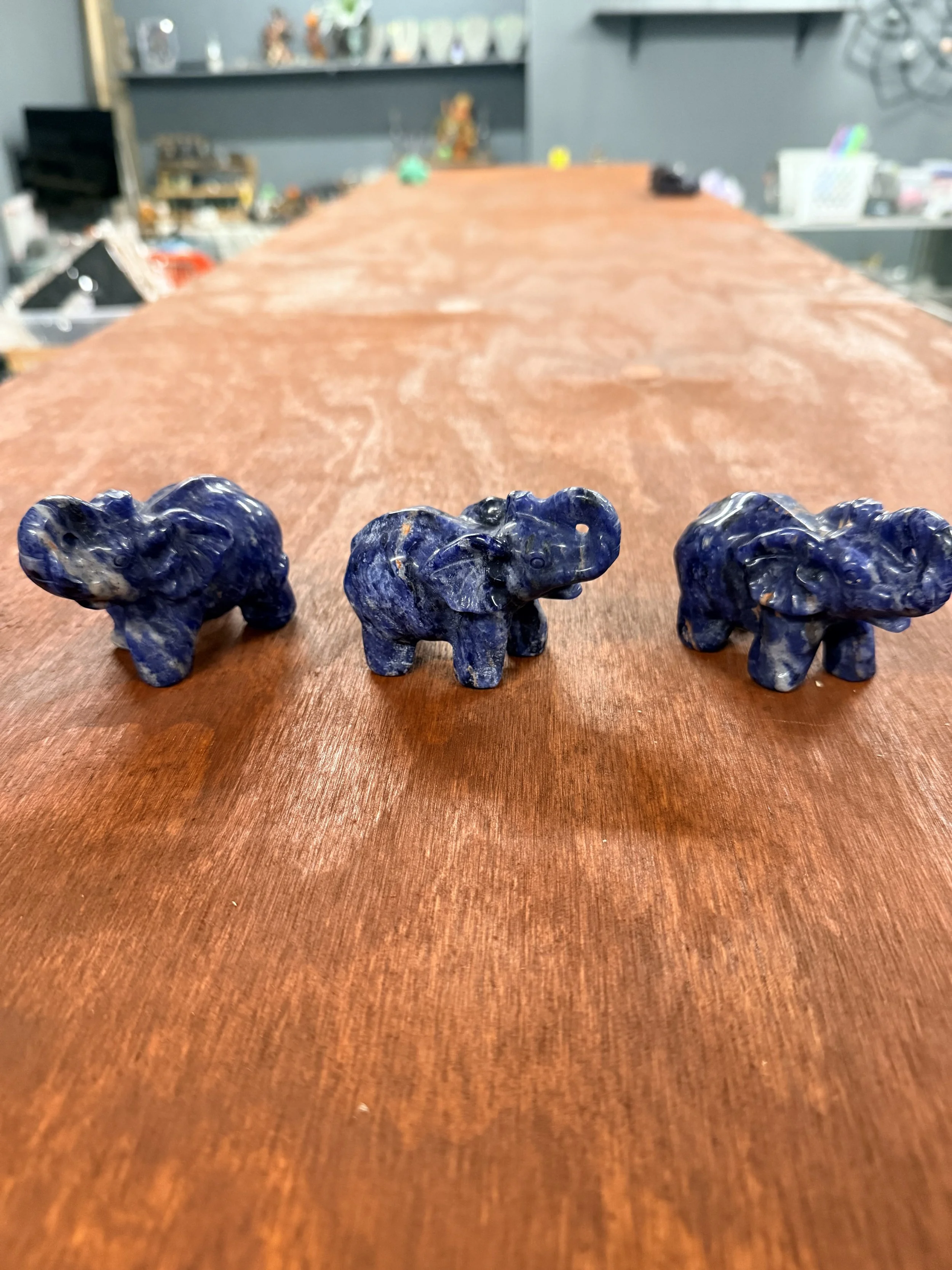 Three blue ceramic elephant figurines on a polished wooden table in a room with various decorations and shelves in the background.