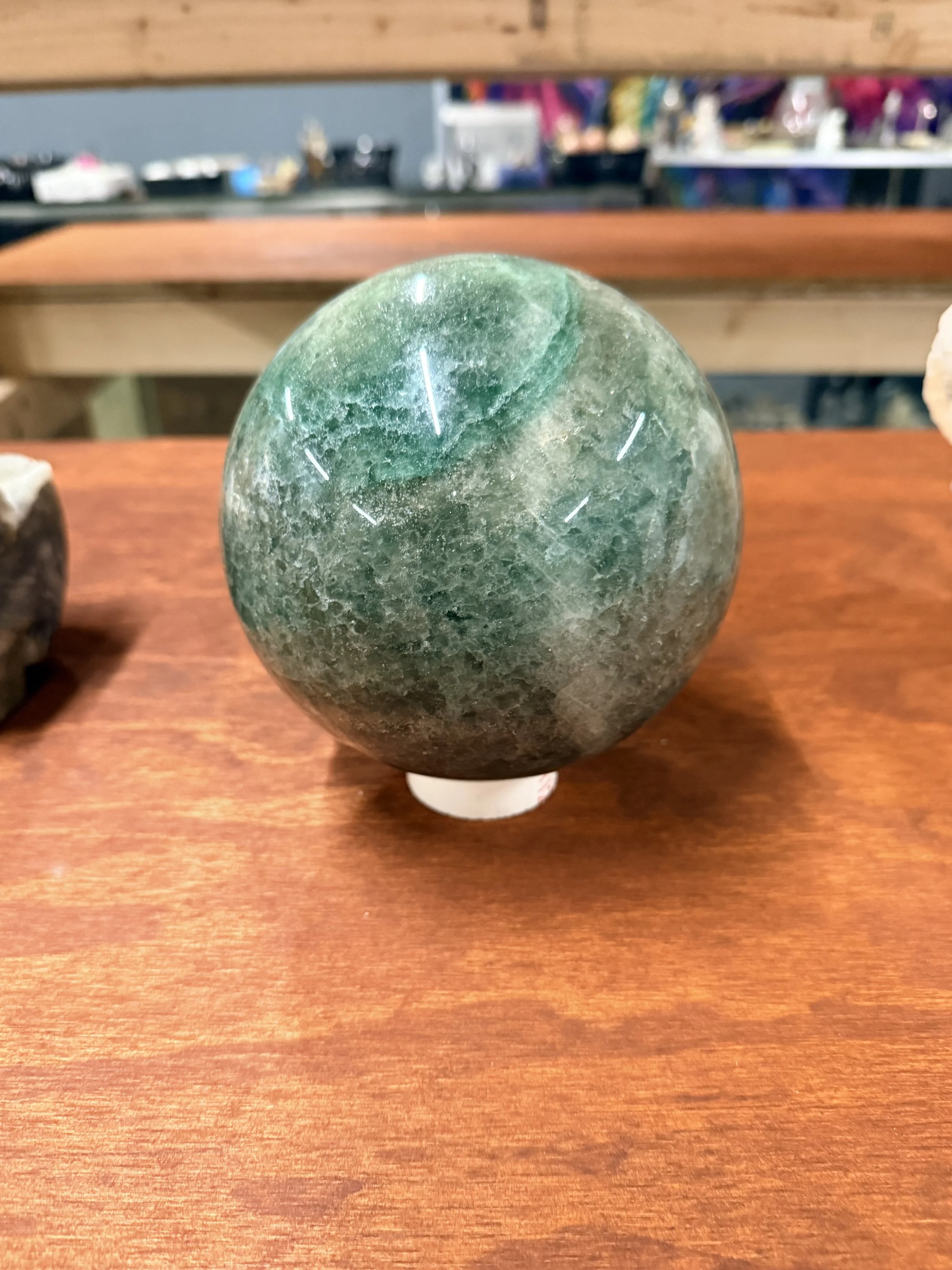 Green spherical mineral or stone with faint white markings displayed on a wooden shelf in a store.