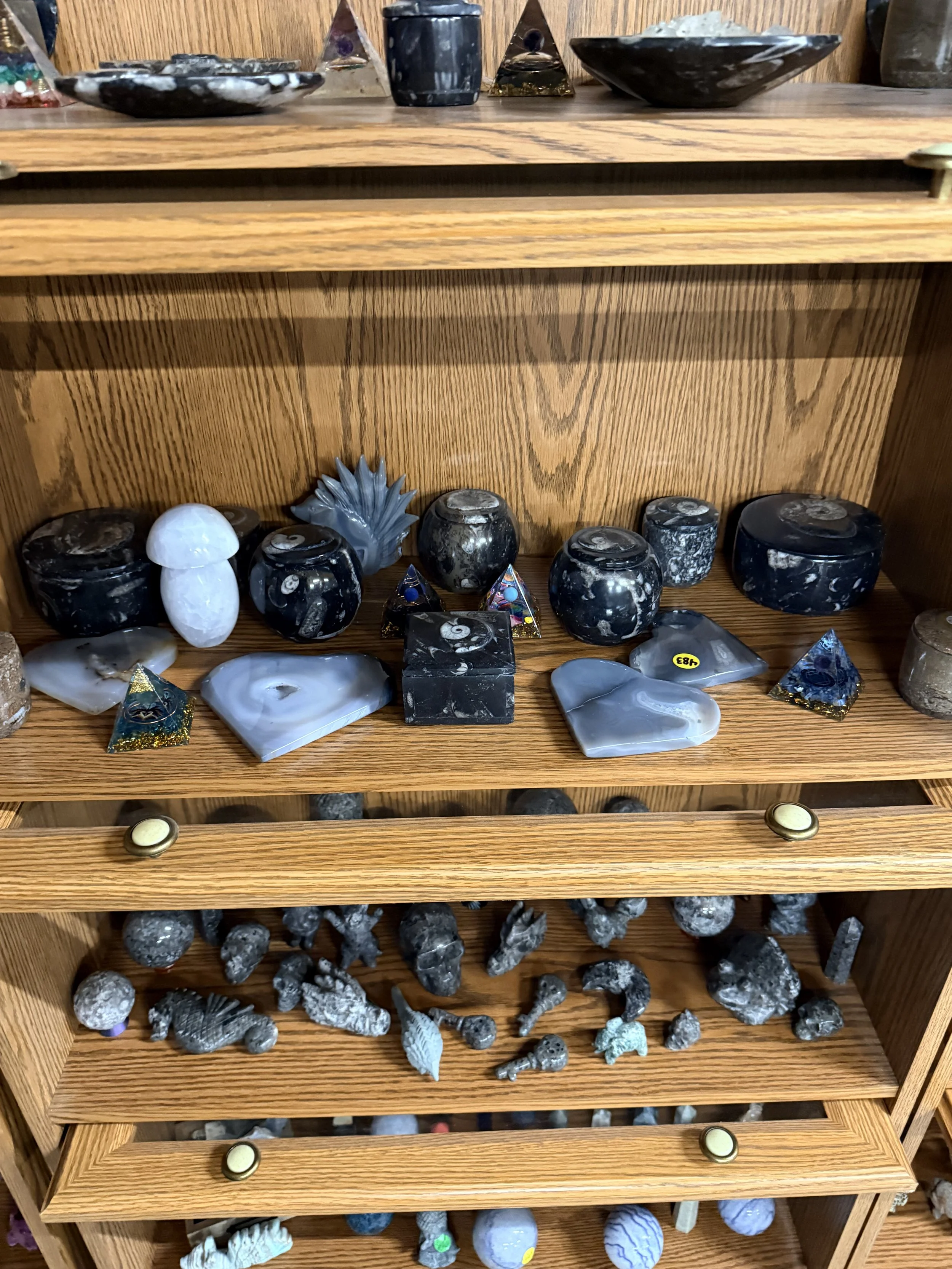 Wooden display shelf holding various black, white, and gray stones and decorative objects, including some with iridescent and metallic accents, arranged on three shelves.