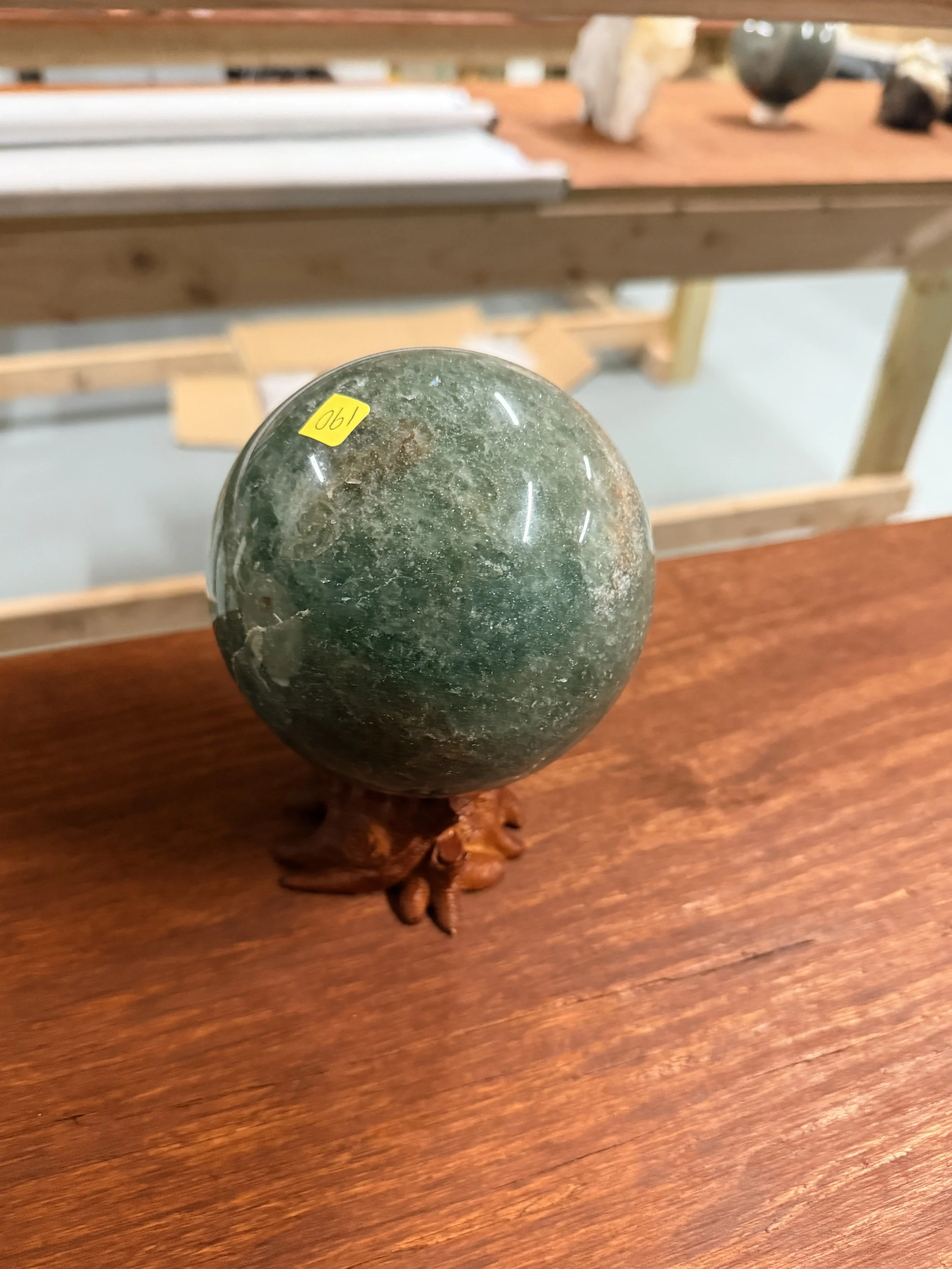 An oval polished green stone on a wooden surface with a small yellow sticker on it that has the number 190 written on it. The background shows a wooden shelf with other stones and objects.