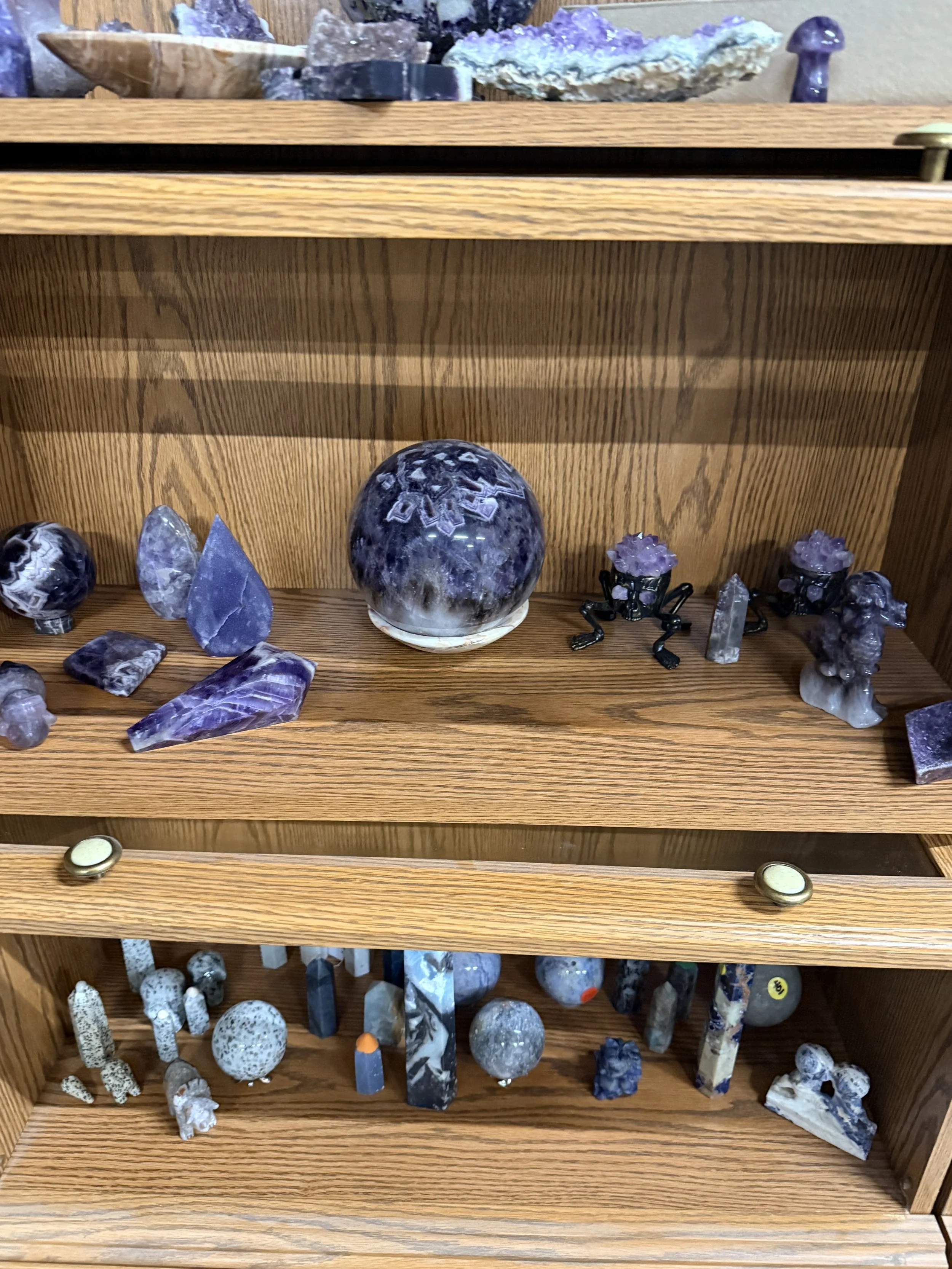 Display of polished and rough gemstones, minerals, and crystals on wooden shelves, including amethyst, quartz, and other semi-precious stones.