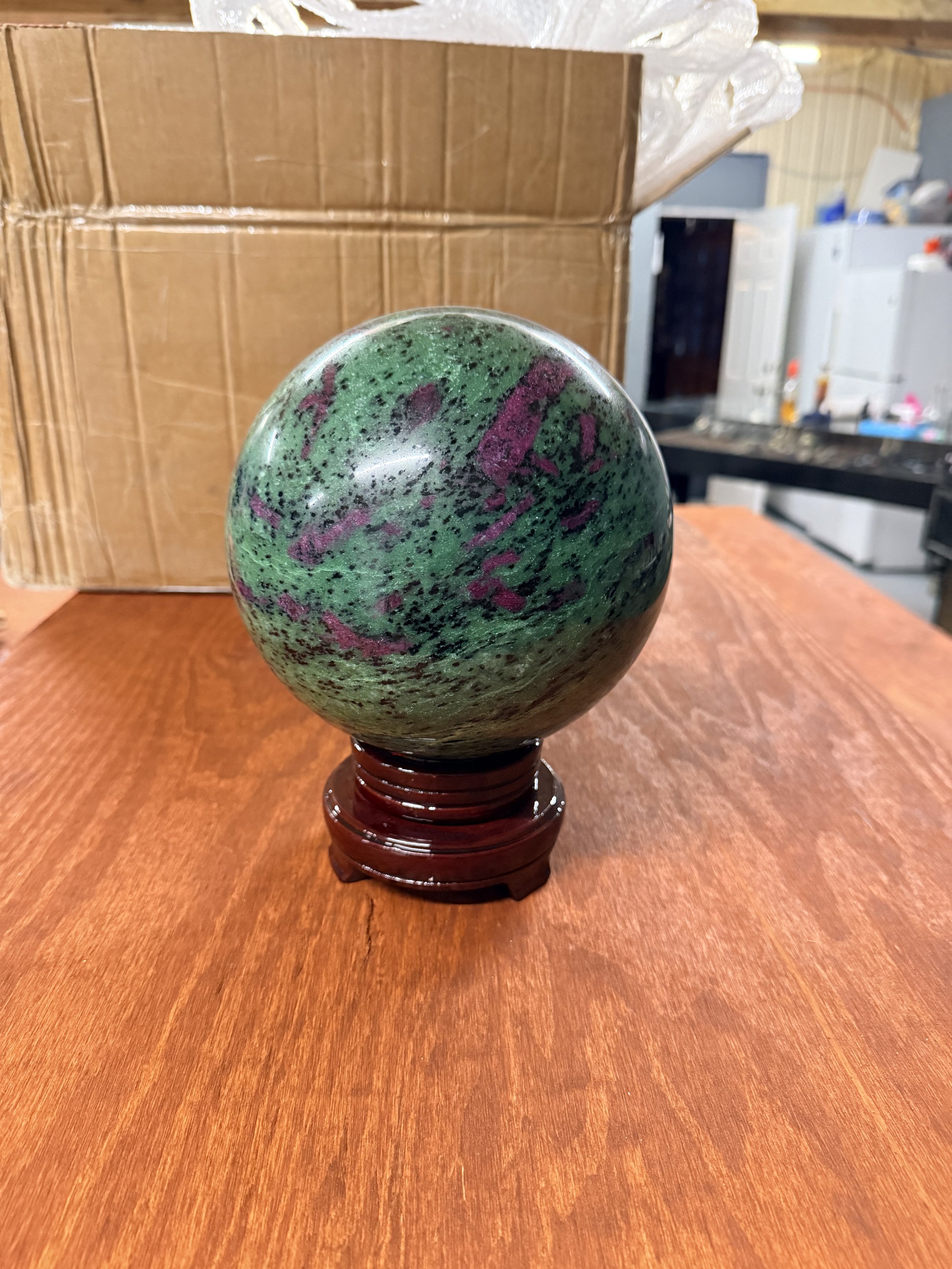 A polished stone globe with green, black, and purple colors sitting on a wooden stand, placed on a wooden table in a room with cardboard boxes and appliances in the background.