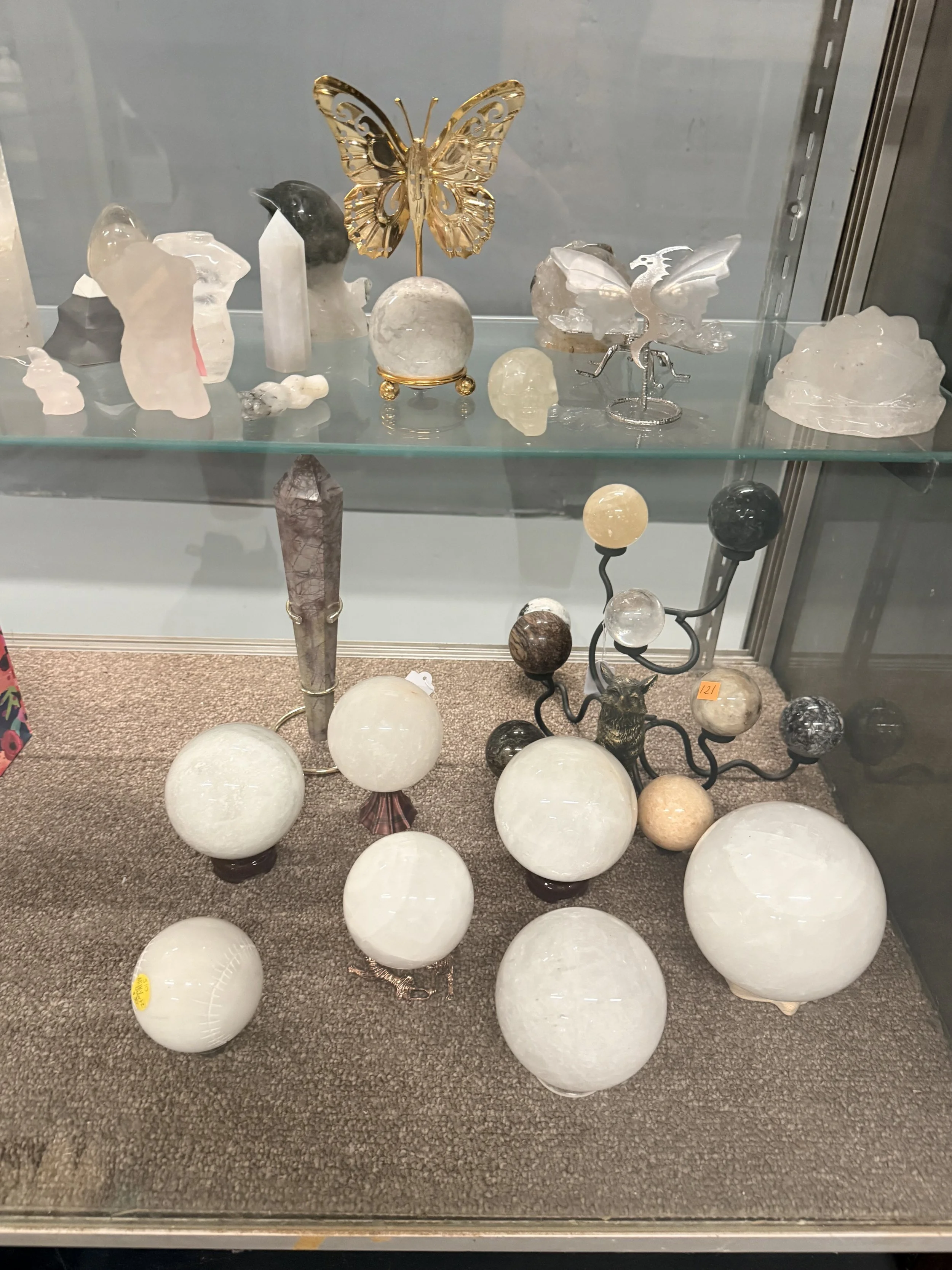 Display of various decorative glass and stone objects, including a gold butterfly ornament, a dragon-shaped sculpture, and multiple spherical and irregularly shaped stones, on glass shelves in a display case.
