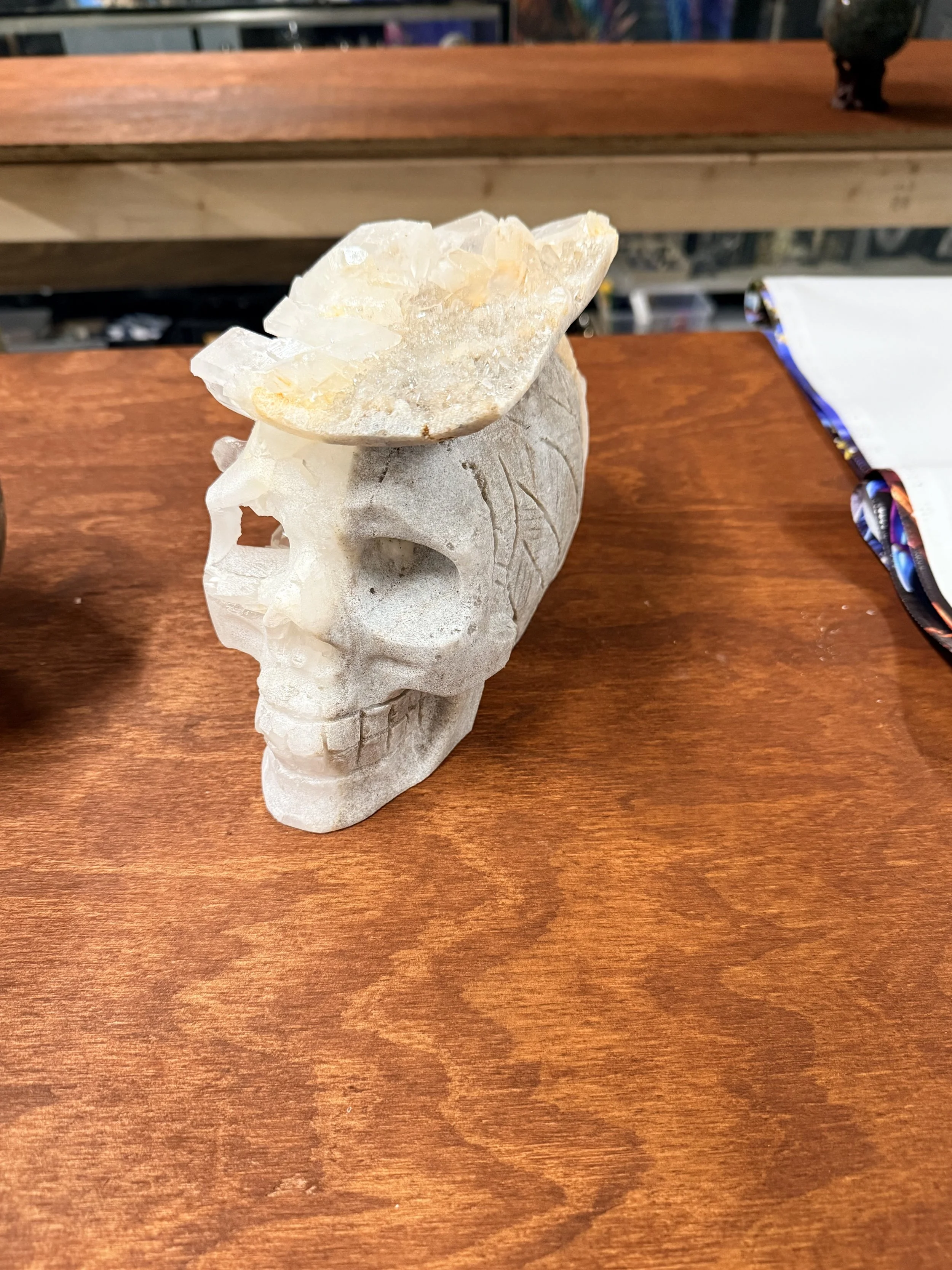 A carved skull sculpture made of white stone with a cluster of translucent, pale yellow mineral crystals on top, placed on a wooden surface.
