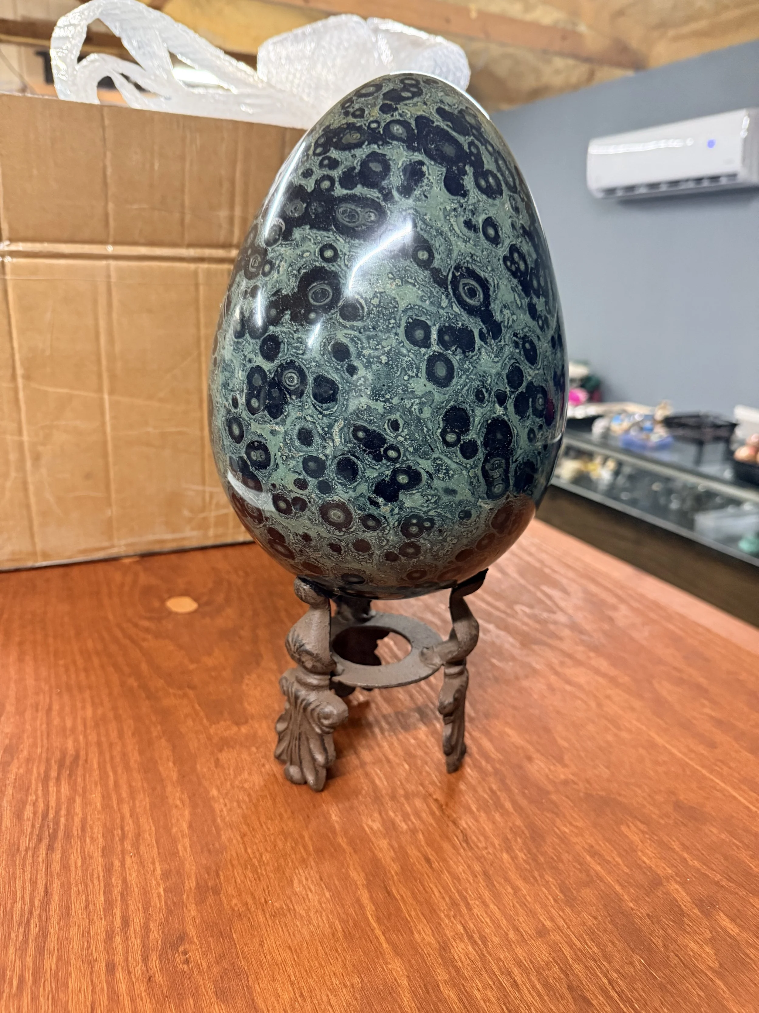 A polished, egg-shaped black and green stone with circular patterns, mounted on an ornate, carved metal stand, on a wooden surface.