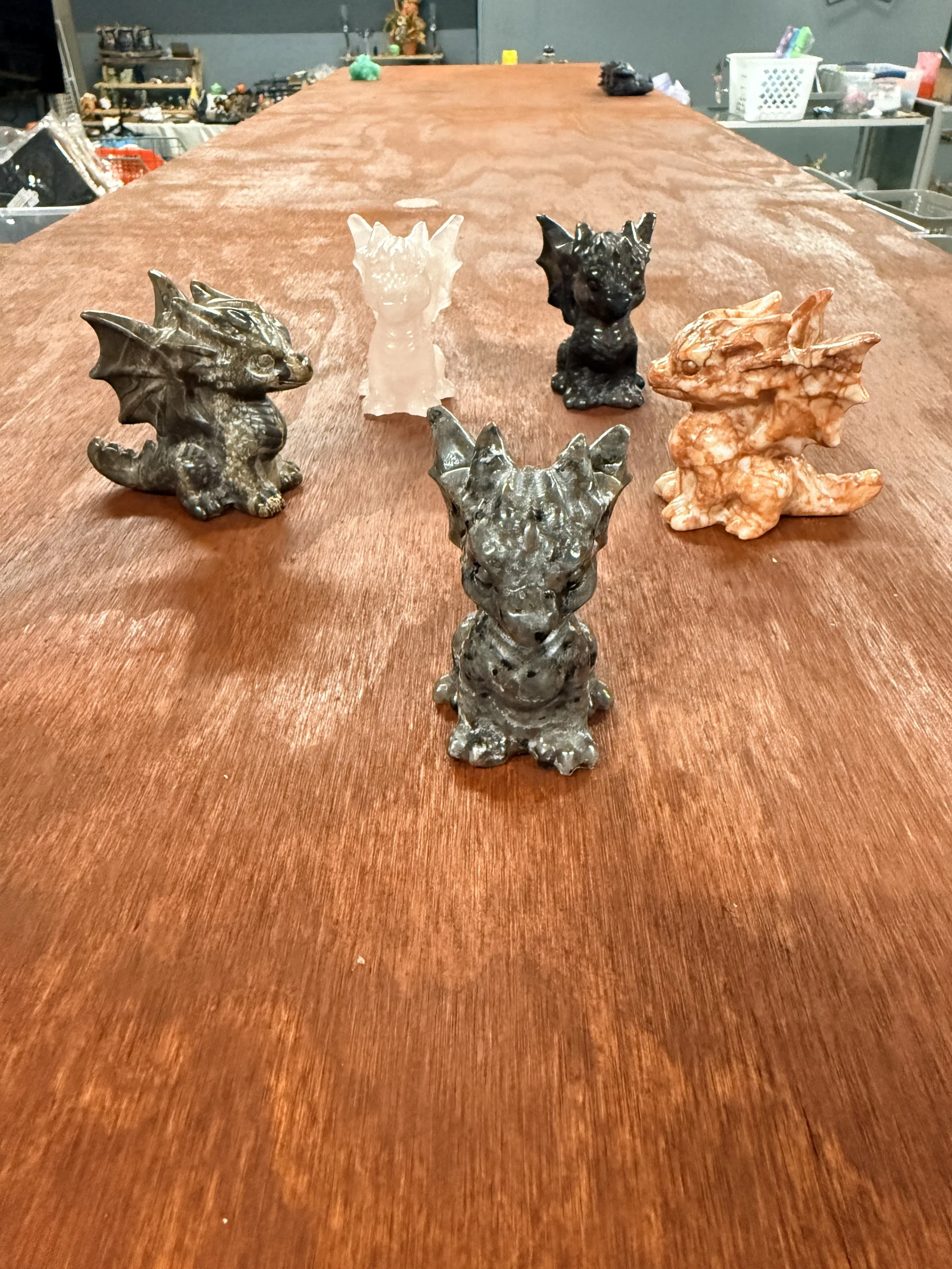 Five small dragon or monster figurines on a wooden surface, with scattered items and a workshop or storage room in the background.