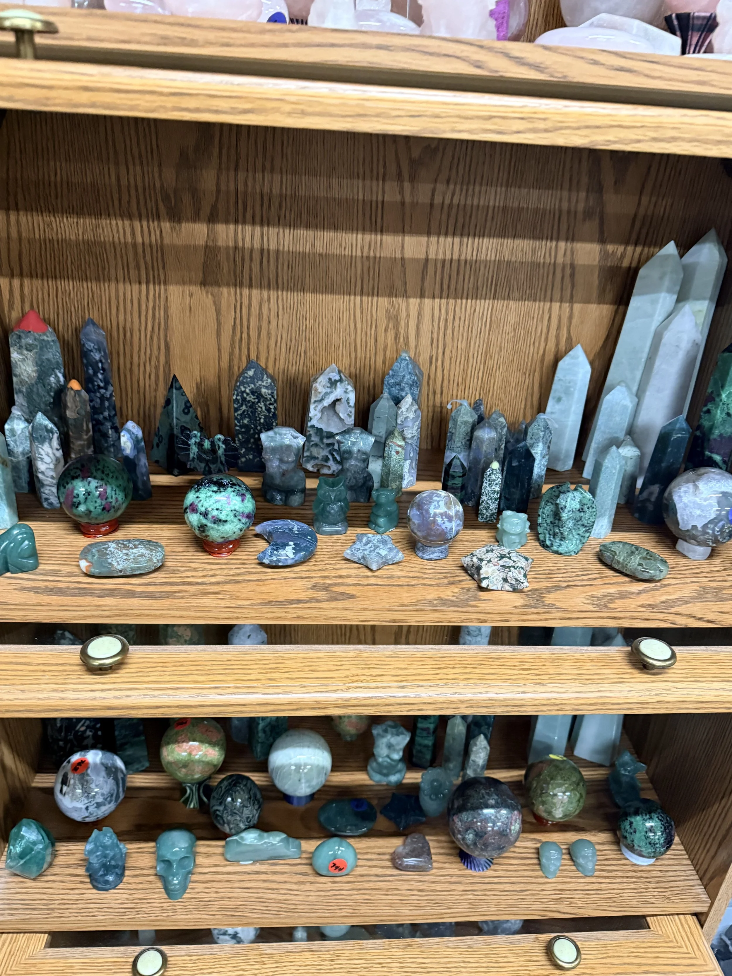 Display case with various carved gemstone figurines and objects, including skulls, spheres, and towers, arranged on wooden shelves.