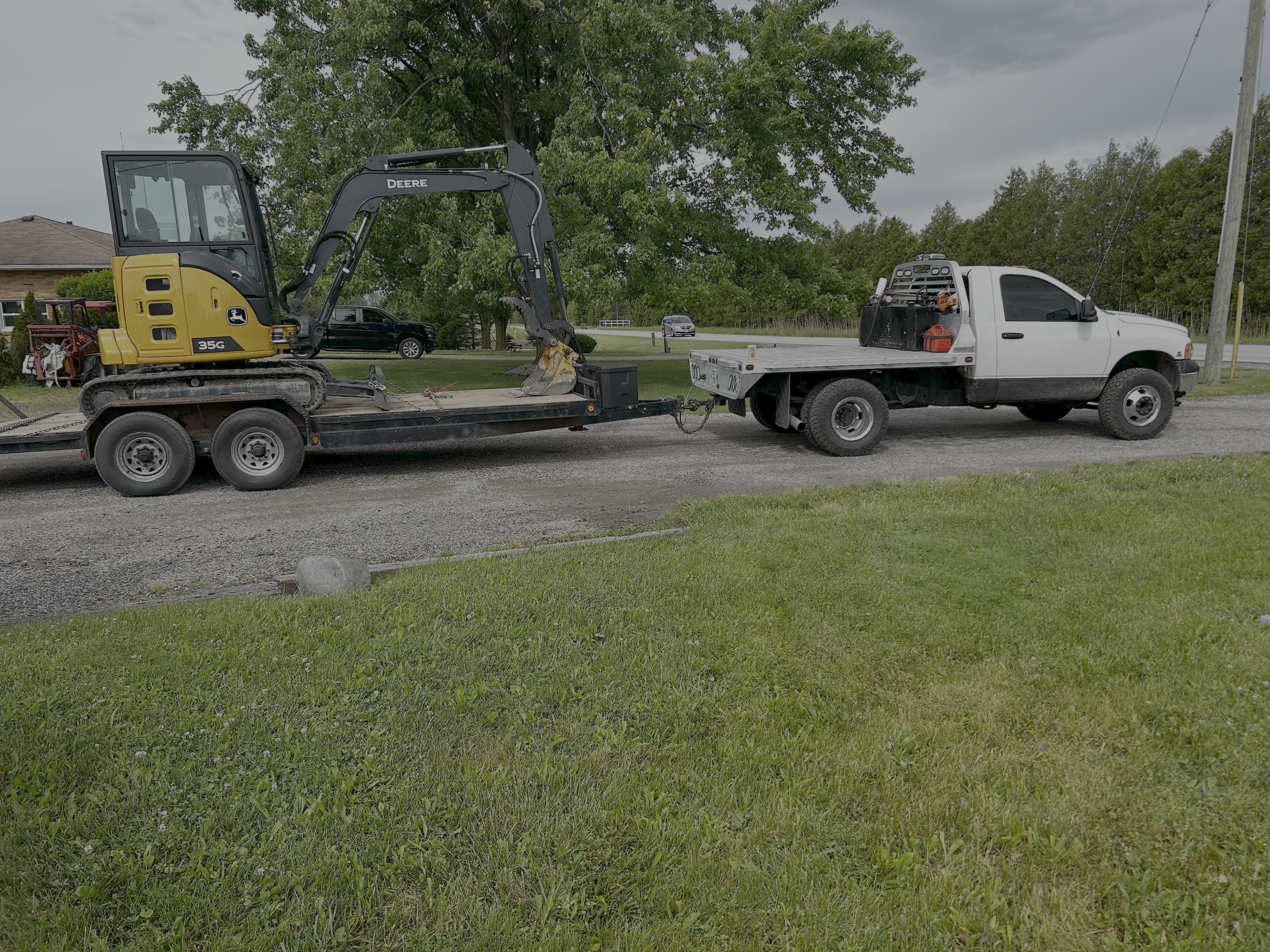 A small John Deere mini excavator on a flatbed trailer hitched to a white pickup truck, parked on a suburban street with trees, grass, and houses in the background.