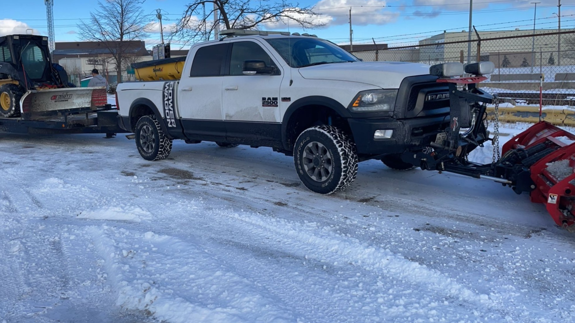 A white Dodge RAM 2500 pickup truck with black accents, parked on snow-covered ground, attached to a snowblower on a trailer.