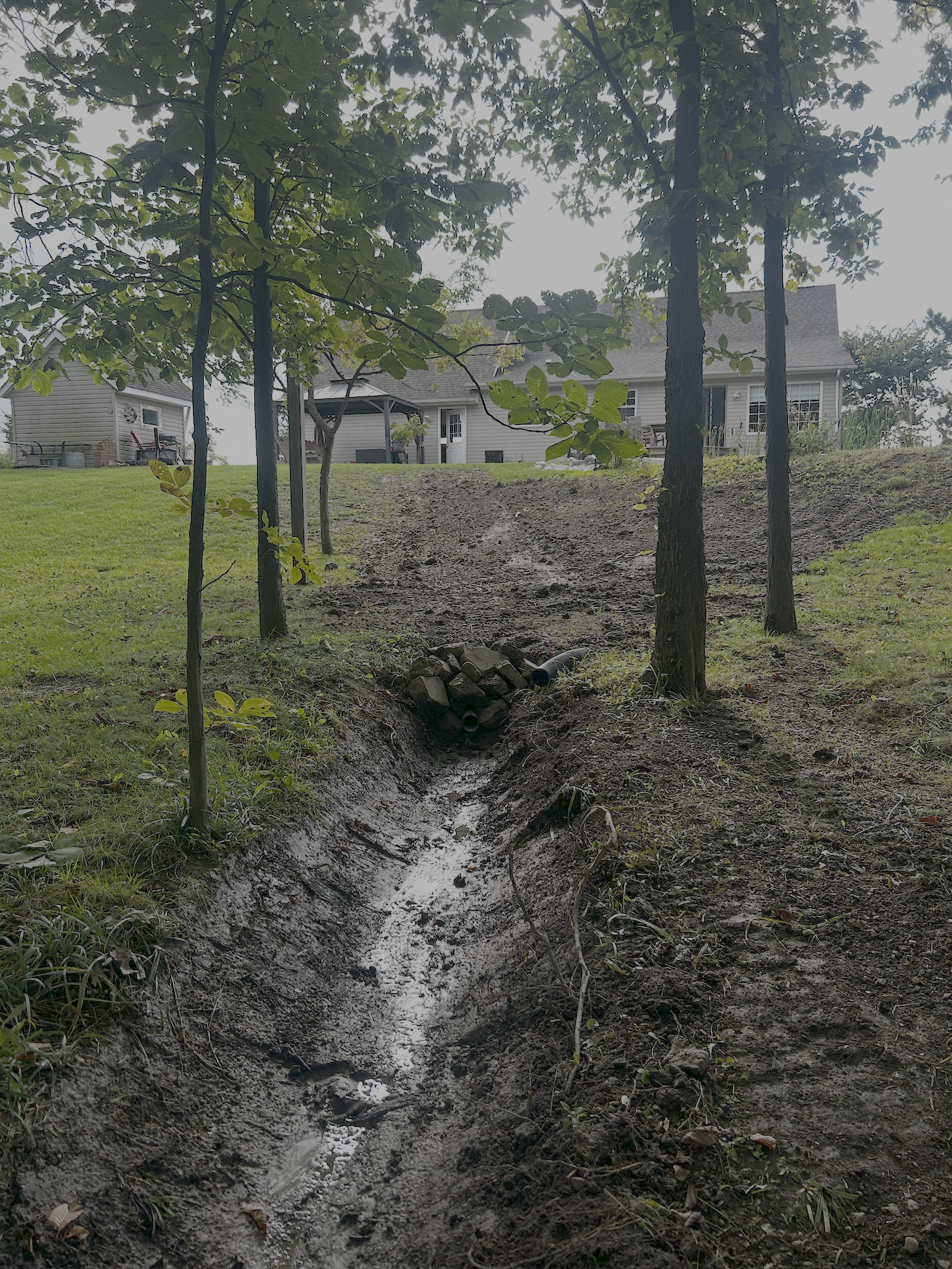 A backyard with a small stream of water flowing through muddy ground, bordered by grass and trees, leading to a house and an outdoor seating area.
