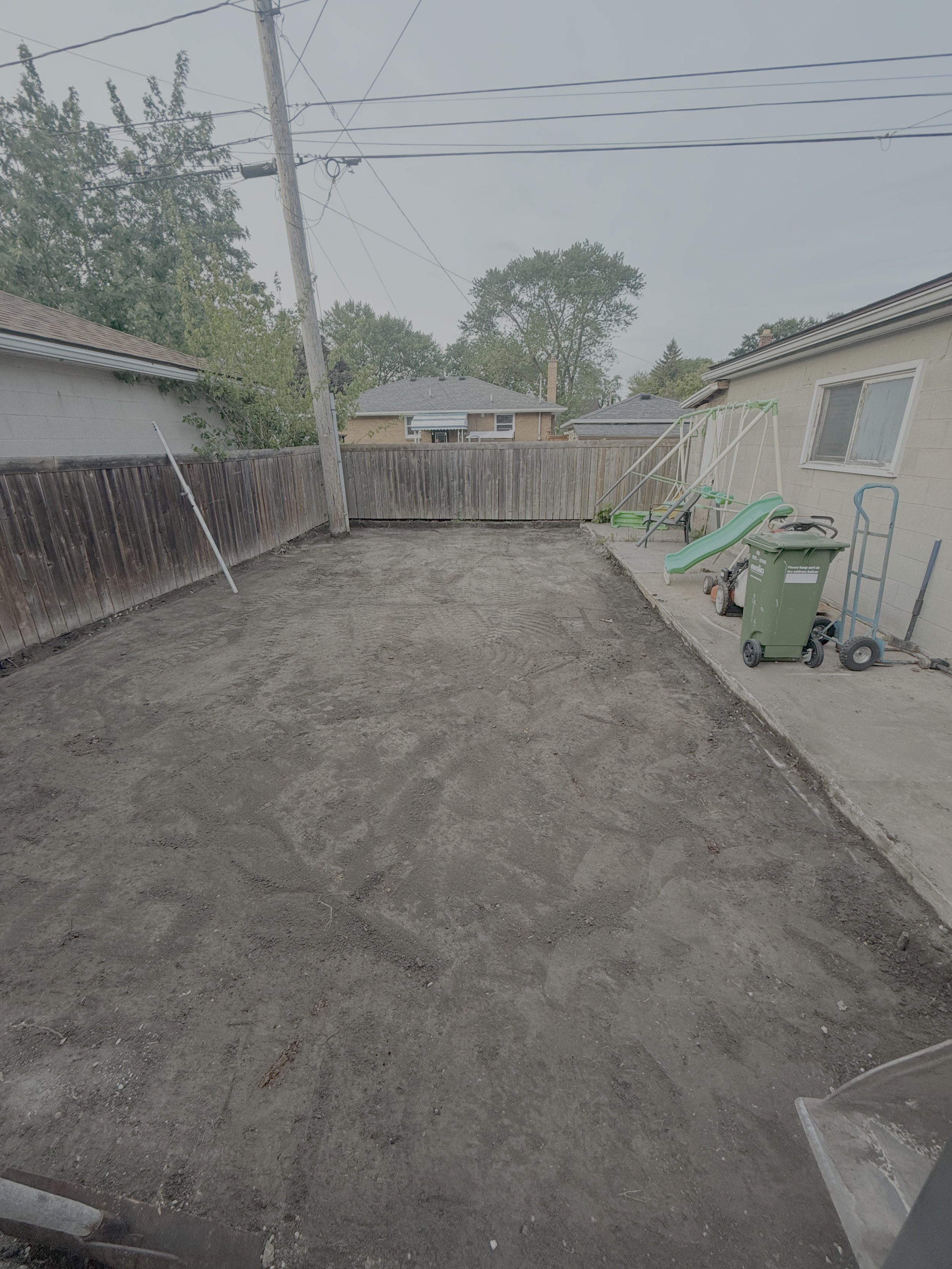 Backyard with a dirt yard, wooden fence, a house with a window, a slide, a swing set, and a trash bin near the house.