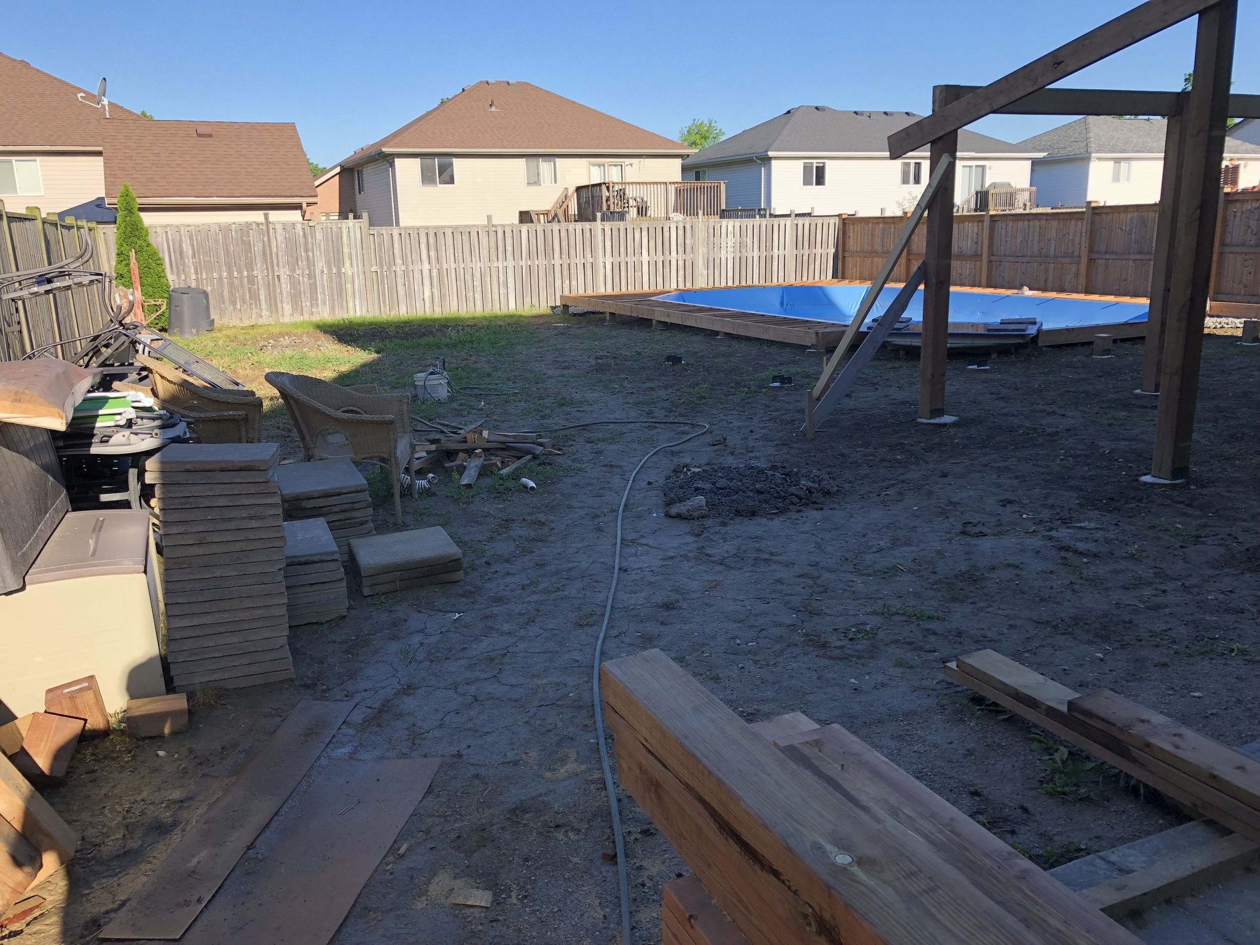A backyard under construction with a partially built above-ground pool, various construction materials, wooden planks, and tools scattered around. The yard is enclosed by a wooden fence, and neighboring houses are visible in the background under a cl
