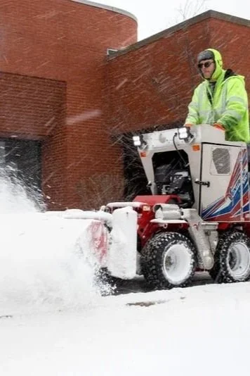 Man in neon yellow jacket operating a snow blower on snowy sidewalk in front of a brick building.