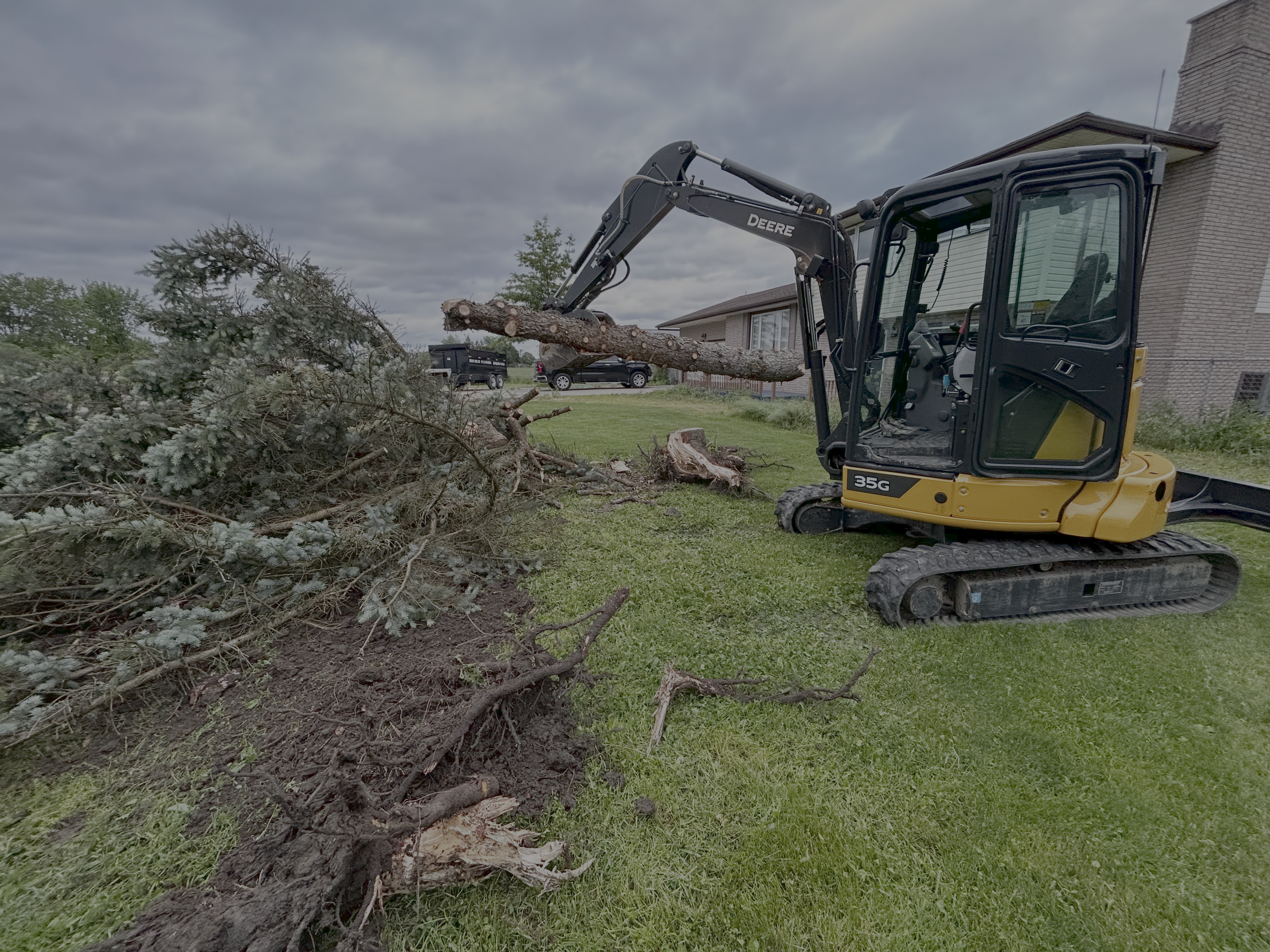 A small John Deere excavator with a black arm is lifting a large fallen tree on a grassy lawn in front of a residential house with a brick chimney. Several parked cars are visible in the background under cloudy skies.