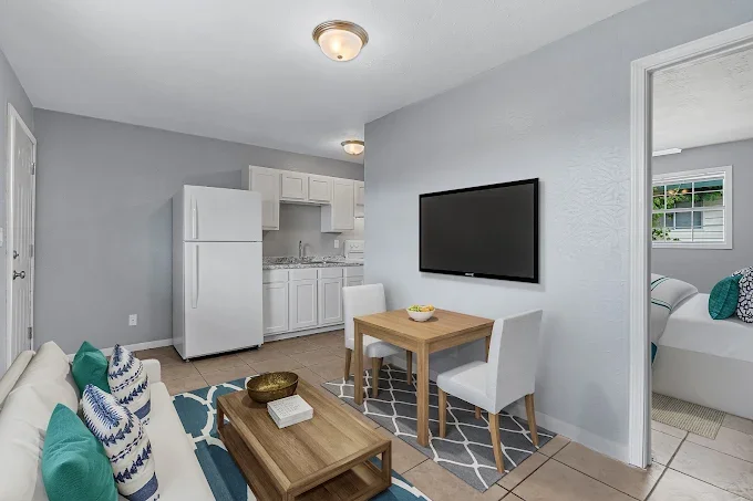 Living area with a white couch, decorative pillows, a coffee table, a wall-mounted TV, a small dining table with two chairs, and a view into a bedroom with a bed and window.