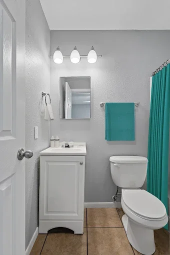 A small bathroom with a white vanity, a mirror, a toilet, a teal towel, a teal shower curtain, and a light fixture with three bulbs.