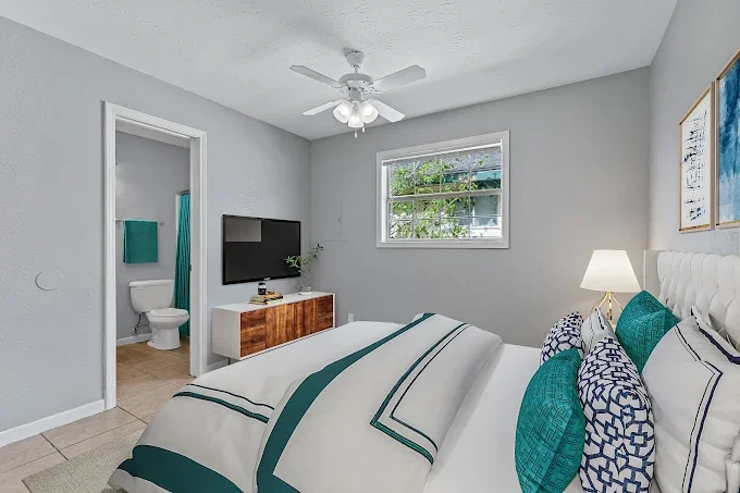 A bedroom with light gray walls, a bed with a white headboard, teal and white bedding, and pillows. There is a nightstand with a lamp, a window overlooking greenery, a ceiling fan with lights, and a flat-screen TV on a wooden dresser. An open door le
