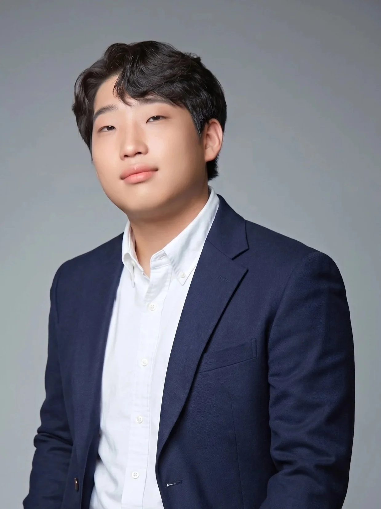 A young man in a navy blazer and white shirt posing against a neutral gray background.