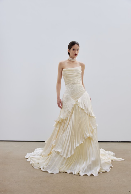 A woman in a strapless, cream-colored, layered gown with pleats and ruffles, wearing a pearl choker necklace, standing against a plain white wall.