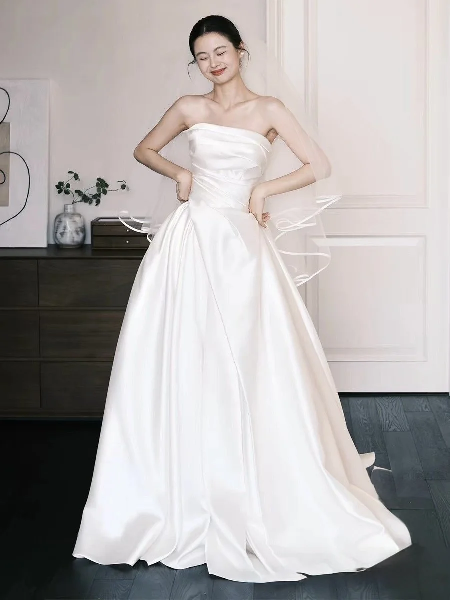 A young woman in a white strapless wedding gown, standing indoors with a happy expression, hands on her hips, surrounded by minimalist decor.