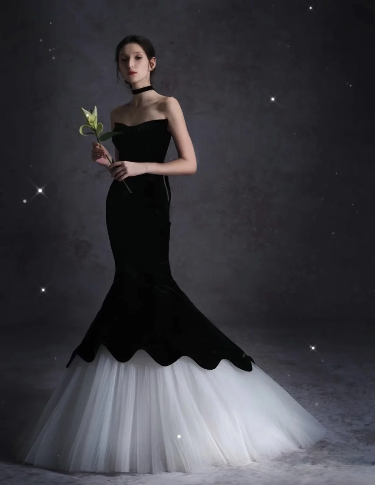 A woman in a long black strapless dress with a white tulle train stands against a starry night sky background, holding a lily flower.