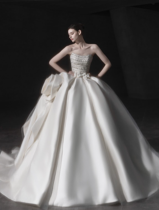 Woman in an elegant strapless white wedding gown with an intricate beaded bodice and a full, voluminous skirt, standing with her hands on her hips.
