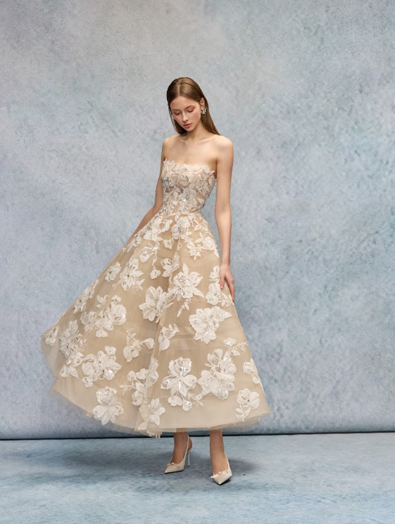 Young woman in an elegant strapless beige dress with white floral lace appliqué, standing against a textured blue background.