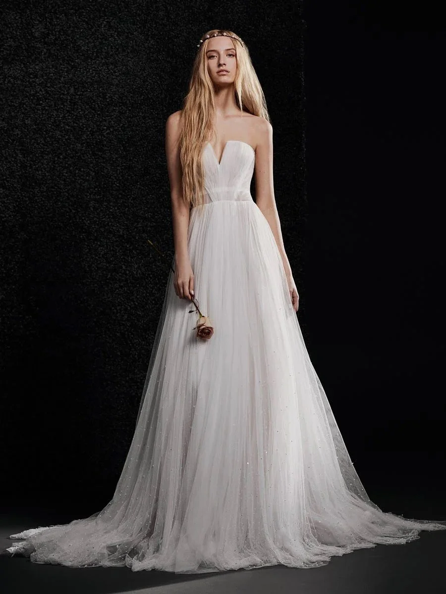 A young woman with long blonde hair in a white strapless wedding dress holds a single pink rose against a black background.