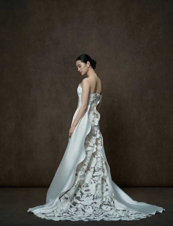 A woman in a strapless wedding dress with lace floral patterns and a satin train, standing against a plain dark background, with her hair styled in an elegant low bun.