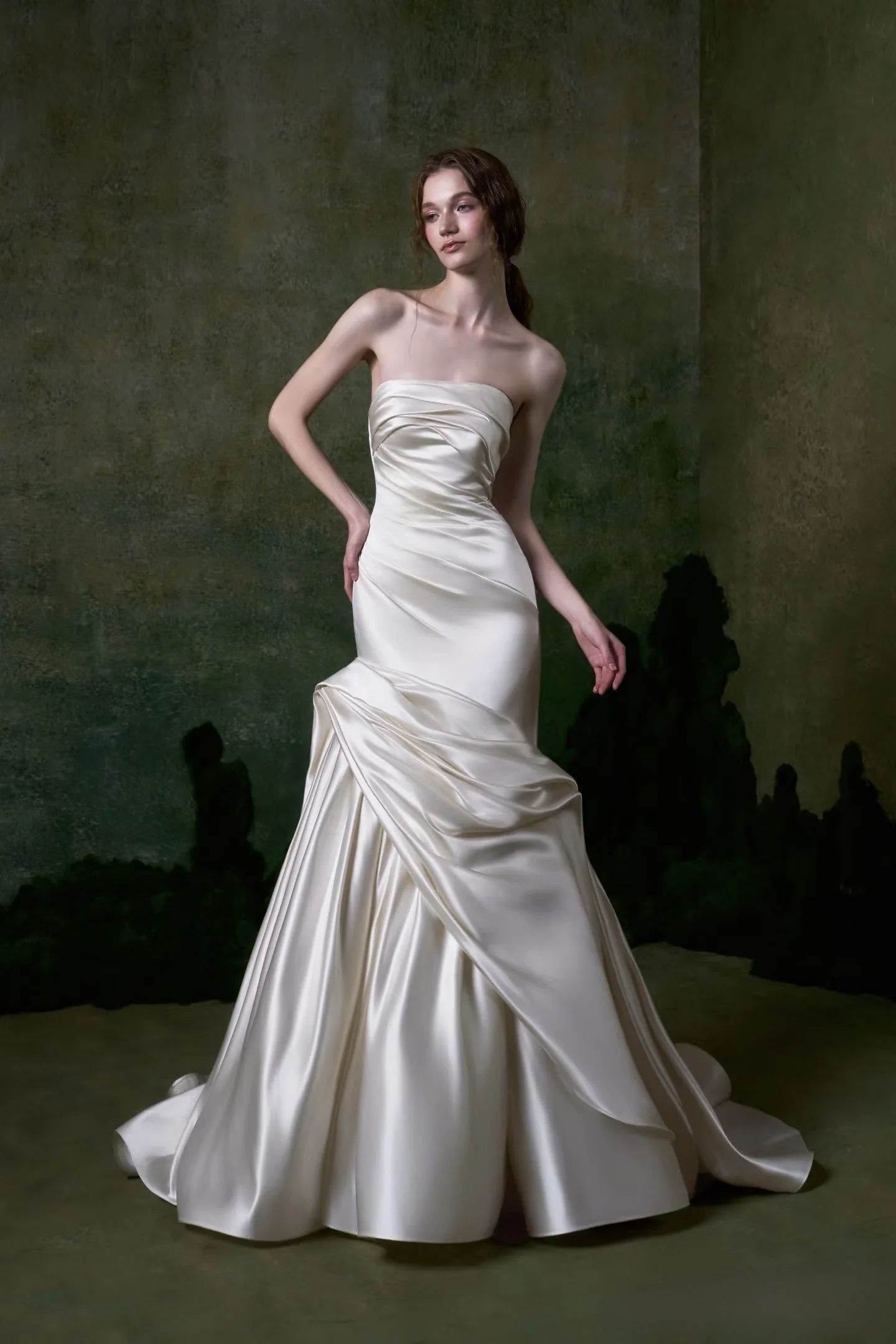 A woman in an elegant, strapless cream-colored gown stands against a dark, textured background. The dress has a fitted bodice with subtle pleats and a full skirt with draped fabric. She has wavy brown hair and a composed expression.