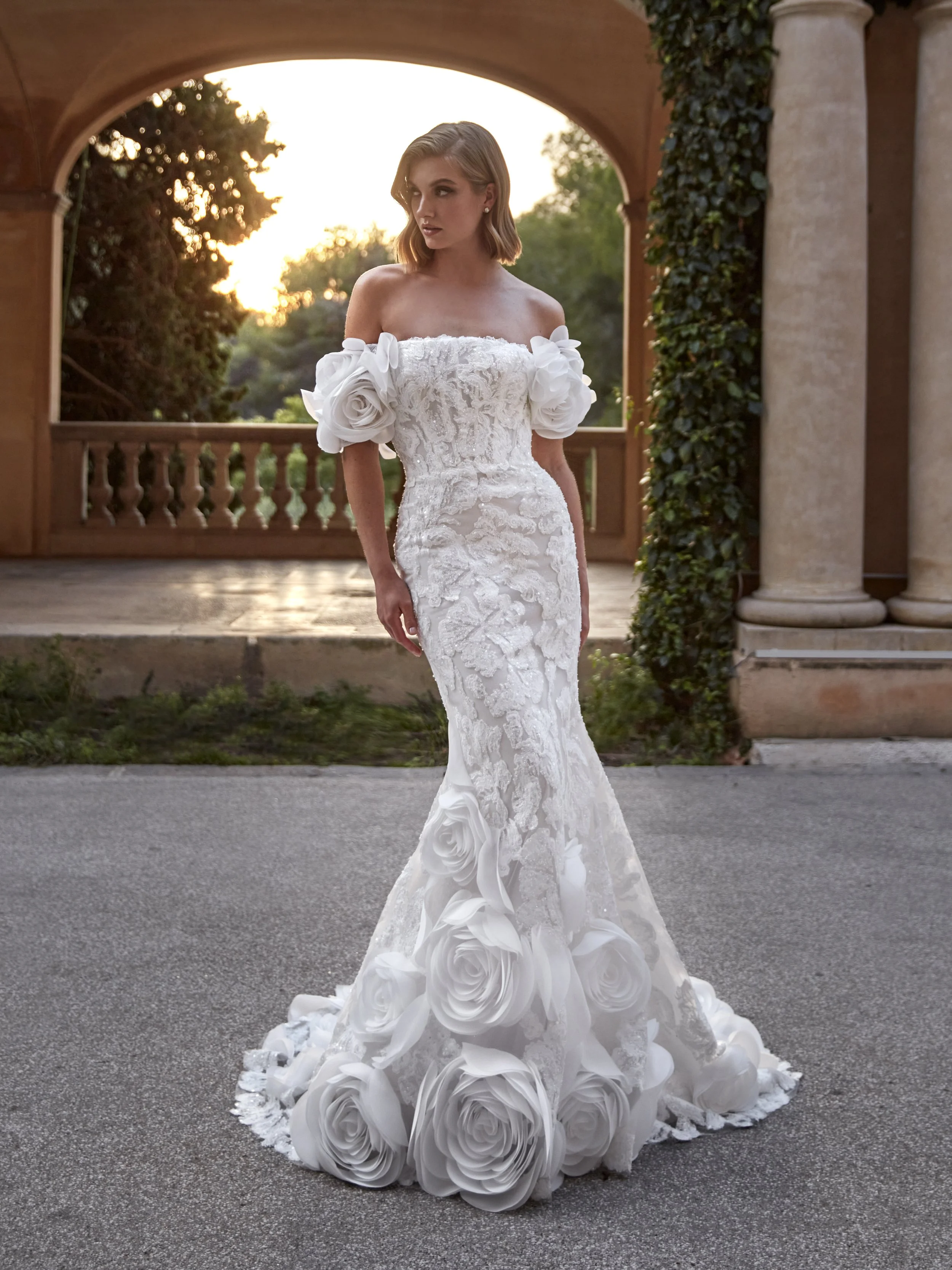 A woman in an elegant white wedding dress with floral details stands outside on a paved surface during sunset, near a stone column covered in ivy, with trees and a balustrade in the background.
