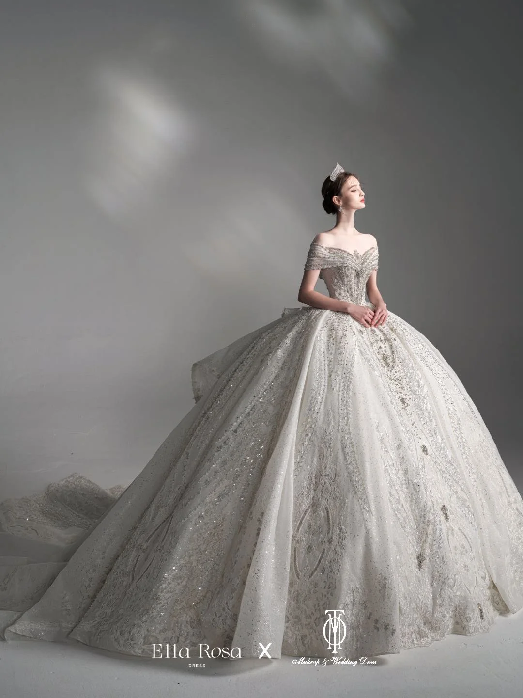A woman wearing an elaborate, off-the-shoulder, silver wedding gown with intricate detailing, standing against a gray background.