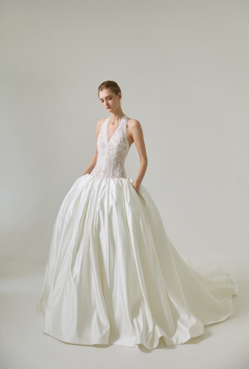 A woman in a white wedding gown with a lace bodice and full satin skirt standing against a plain light gray background.