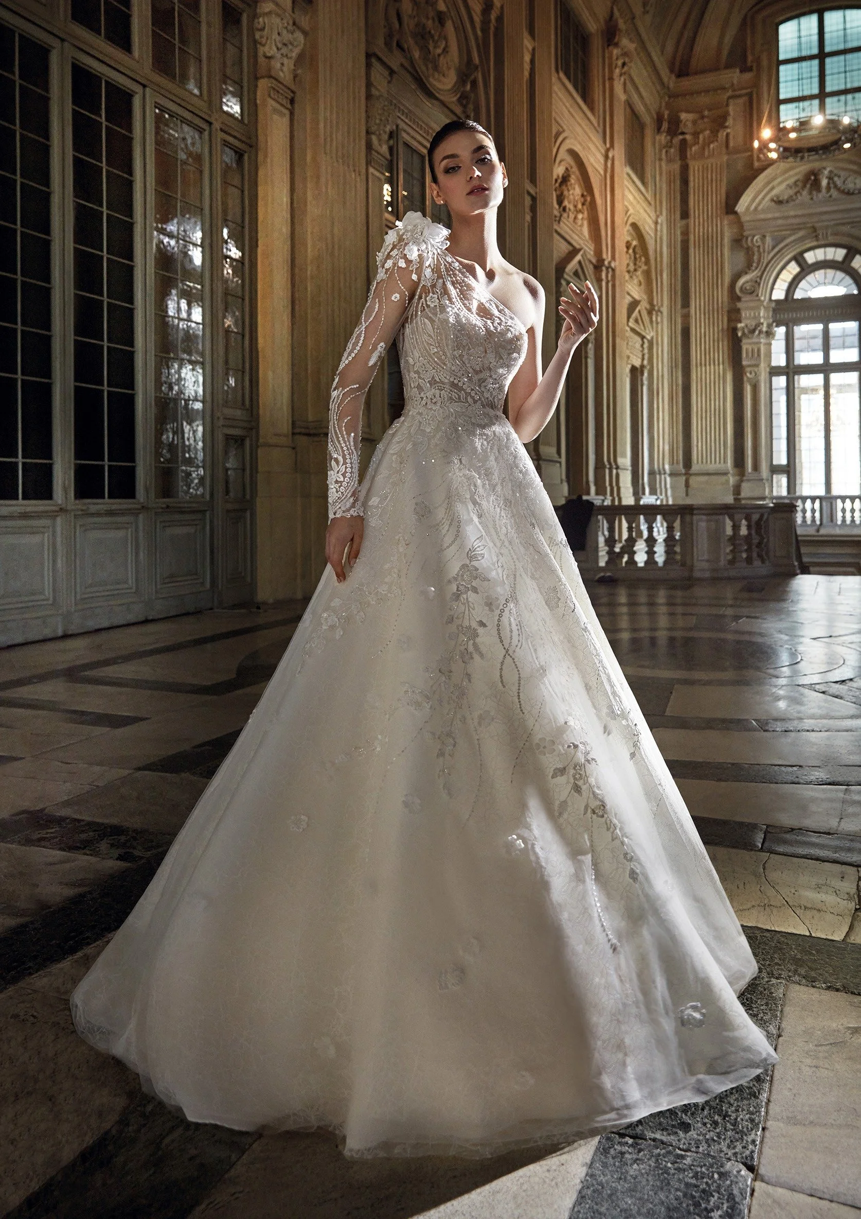 A woman in an ornate wedding gown stands in a grand, historic room with tall windows and detailed woodwork.
