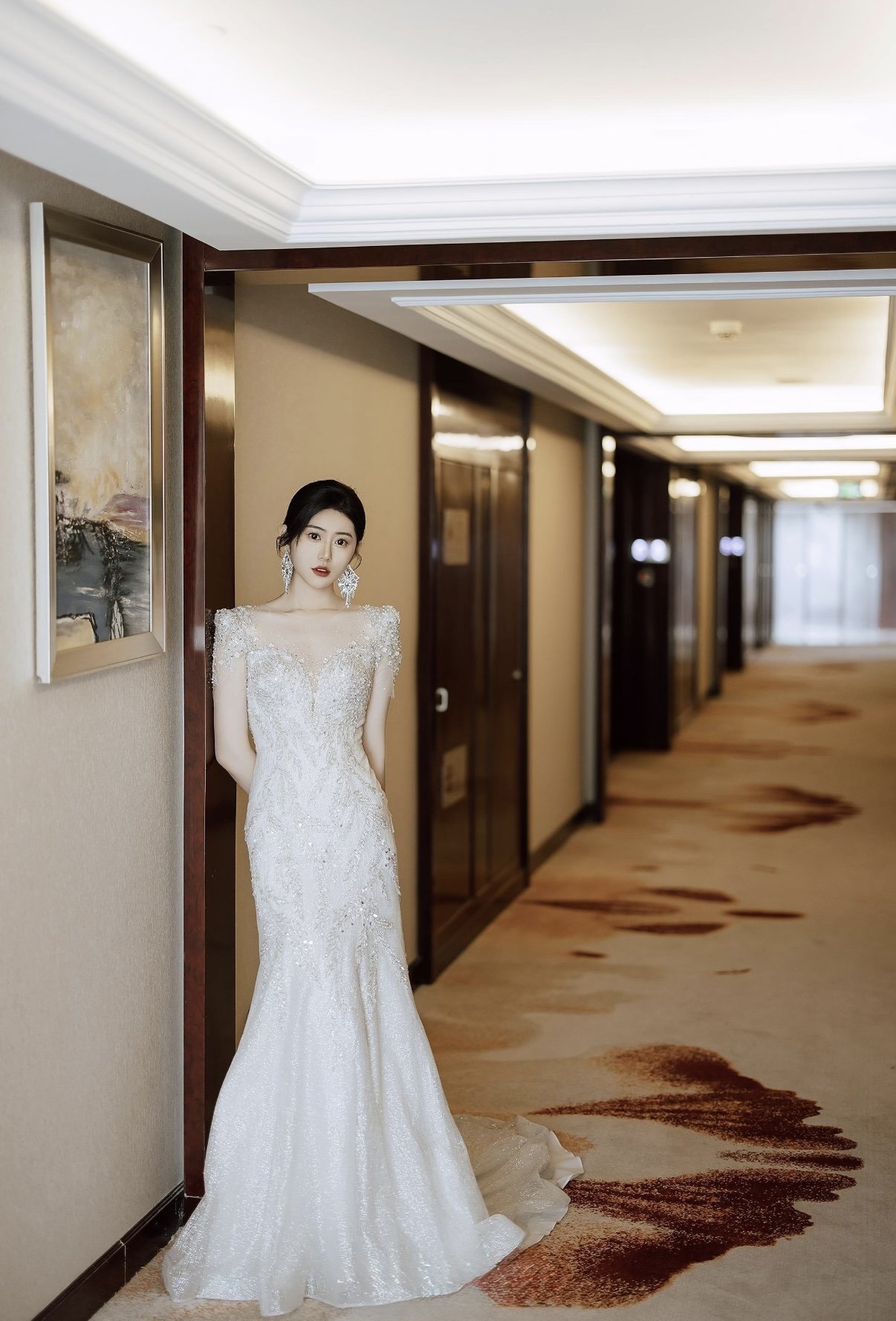 A woman wearing a white wedding gown standing in a hotel corridor near a wall, with her back against it, looking at the camera.
