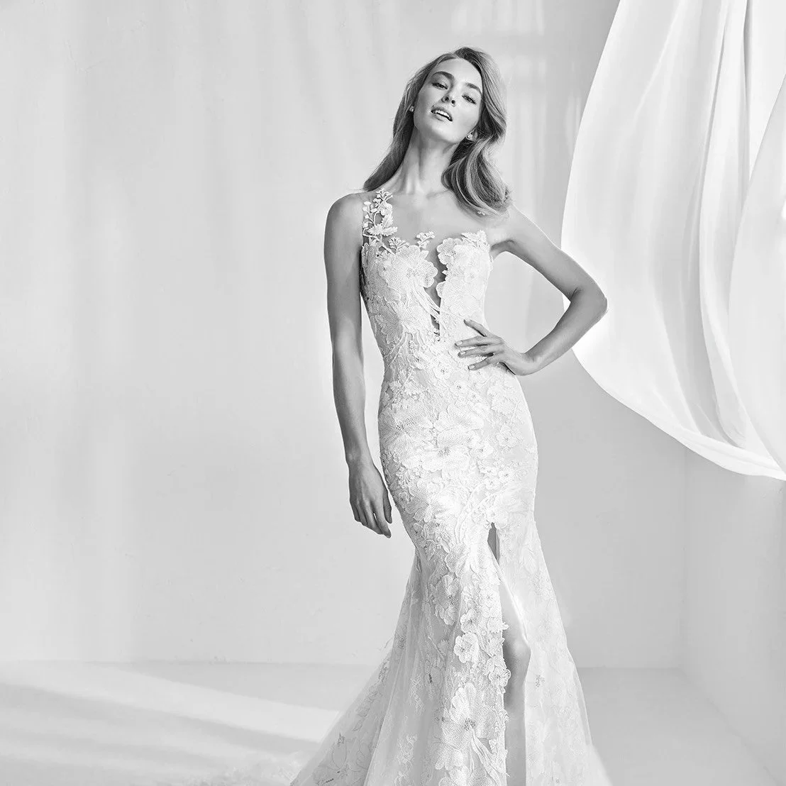 A woman wearing a strapless lace wedding gown with floral appliqués, standing with one hand on her hip in a minimalistic studio setting.