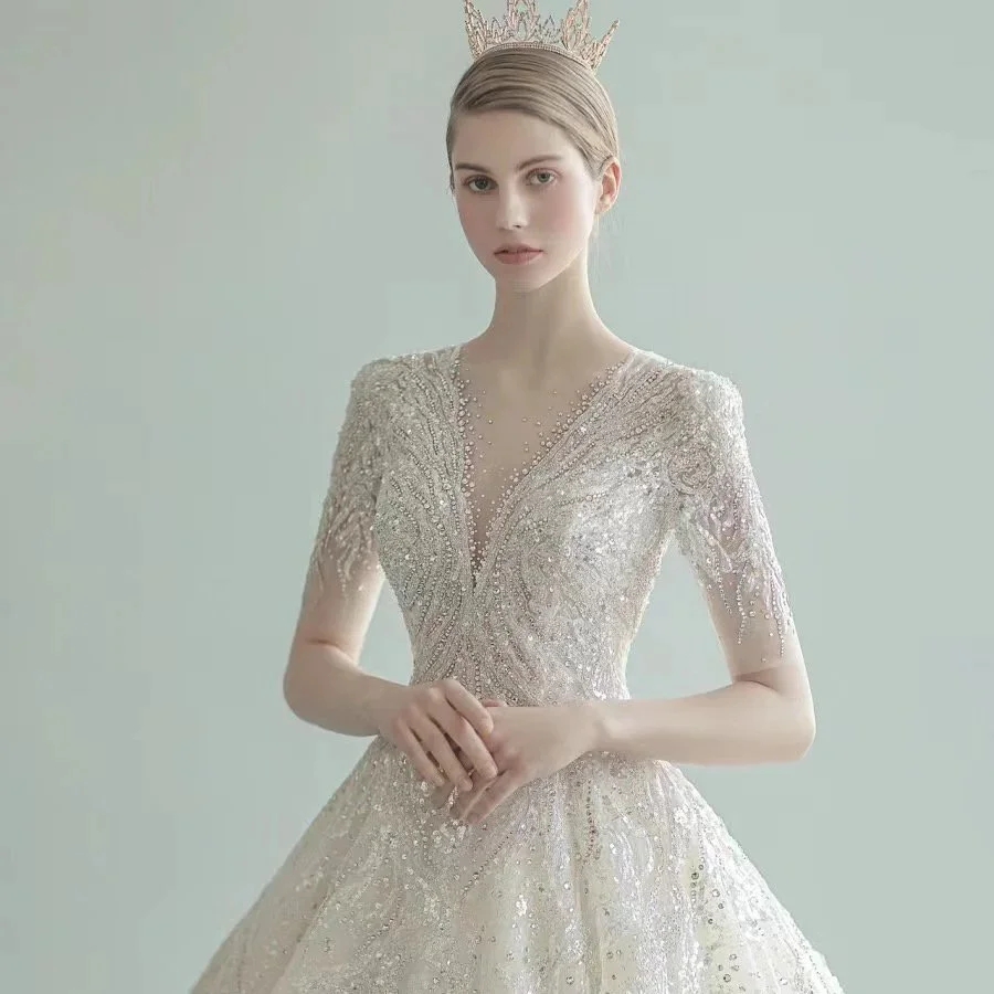 A young woman with fair skin and blonde hair wearing a sparkling, elaborate wedding dress with intricate embroidery and beadwork. She is also wearing a tiara and has a neutral expression.