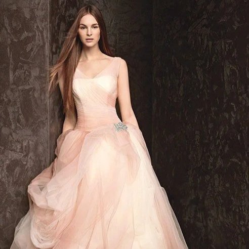A young woman with long brown hair wearing a light pink, strapless ball gown with a fitted bodice and layered tulle skirt, standing against a dark textured background.