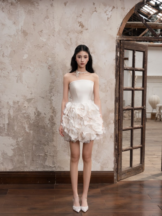 A woman in a white strapless dress with floral detailing, standing indoors in front of a textured wall and an open wooden door.