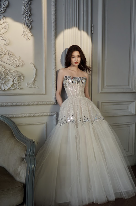 A woman wearing an elegant, strapless, cream-colored gown with sequins and tulle, standing in a decorated room with ornate wall details and a sofa.