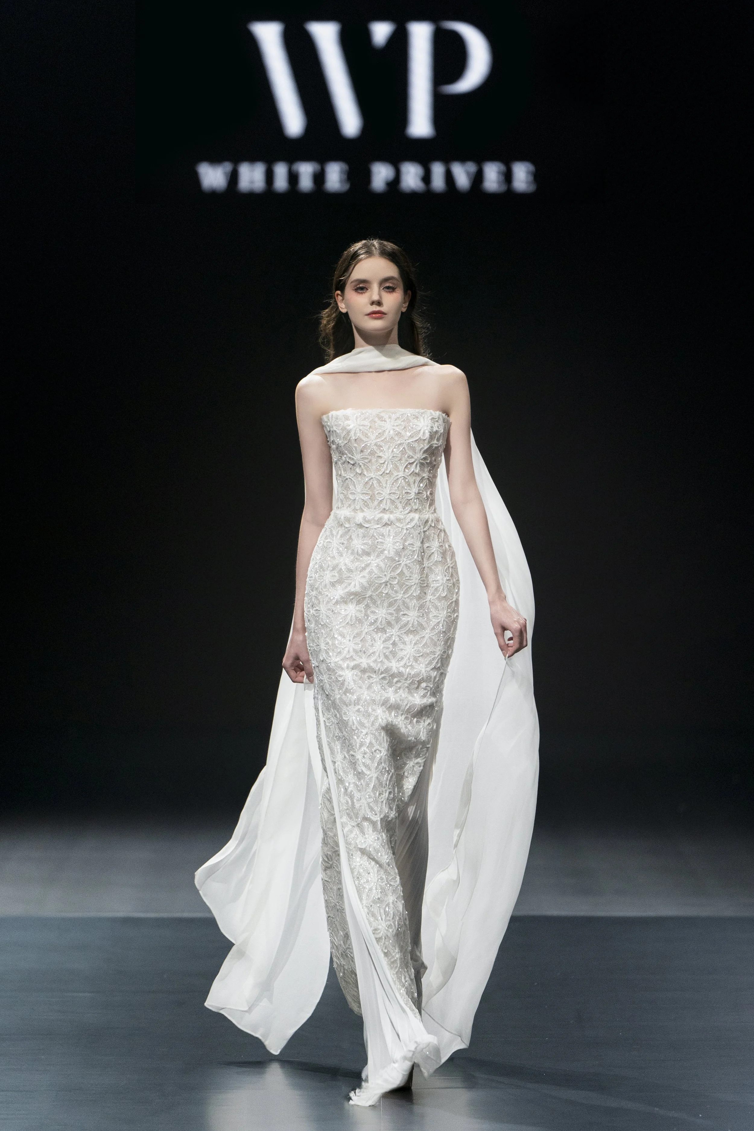Model walking on runway in a white, intricate lace wedding dress with a high slit, flowing train, and a white cape, at a fashion show with a 'White Privé' logo in the background.
