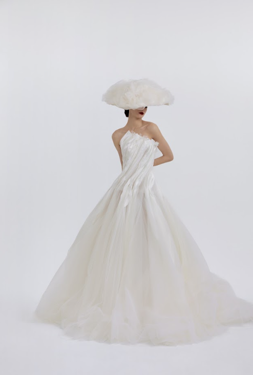 A woman in a white strapless wedding gown with ruffled details, wearing a large fluffy cloud-shaped hat, against a plain white background.
